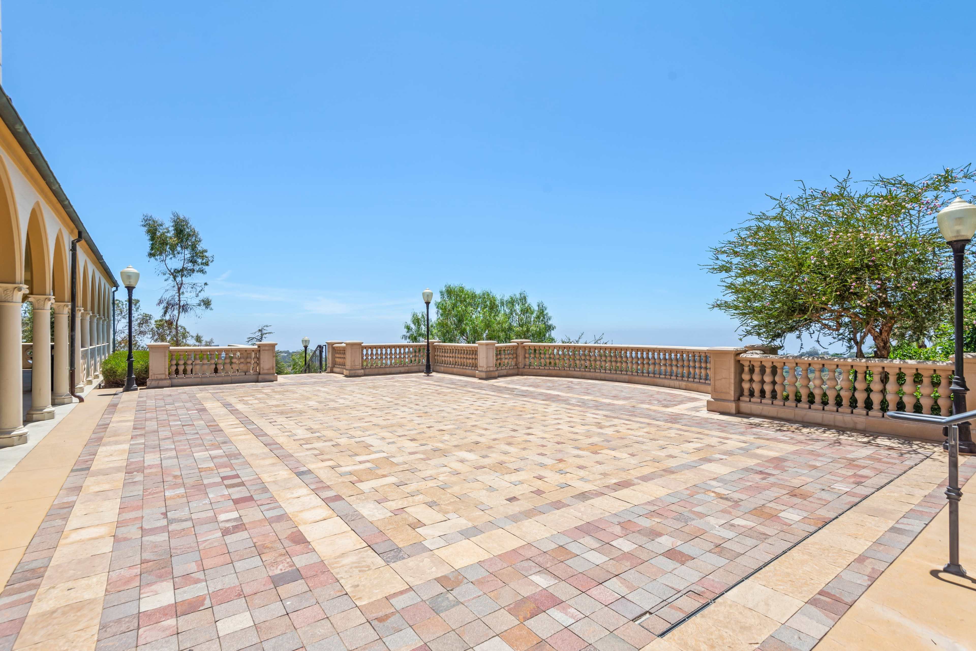 The image shows a spacious stone-paved terrace with decorative railing and trees, overlooking a clear sky and distant landscape.