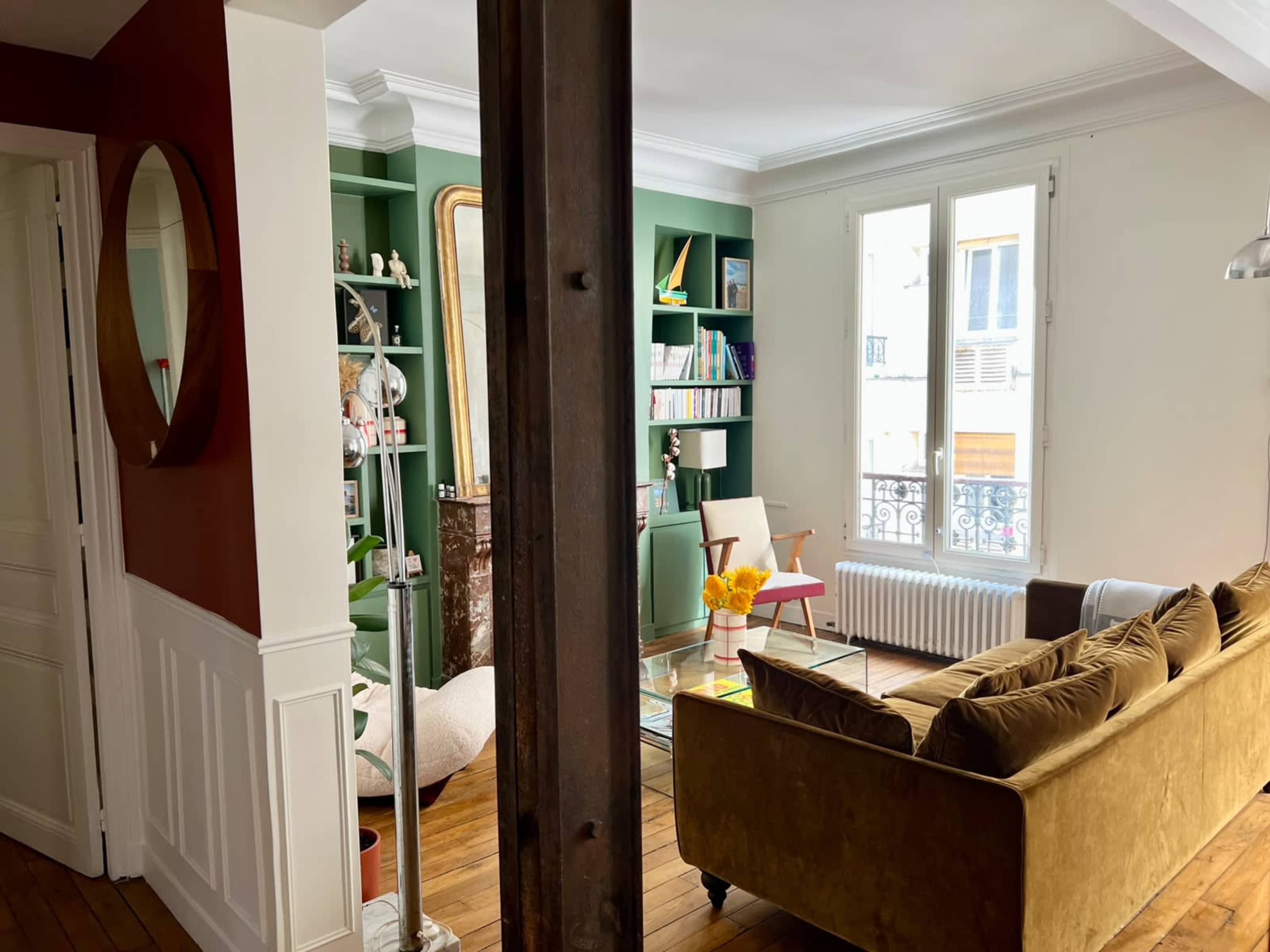 The image shows a cozy living room with a green accent wall, a brown sofa, a bookshelf, and large windows allowing natural light to enter.