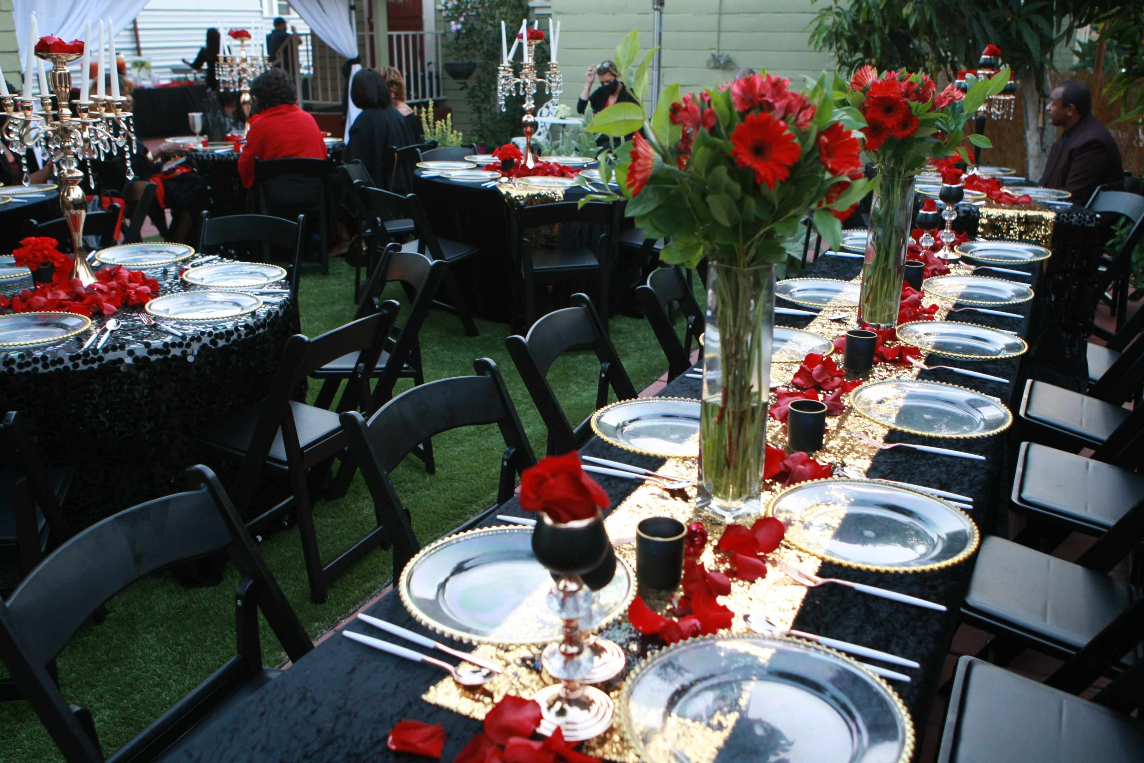 A beautifully arranged outdoor dining setup features black and gold tablecloths adorned with red flowers and rose petals, surrounded by elegantly set tables with black chairs.