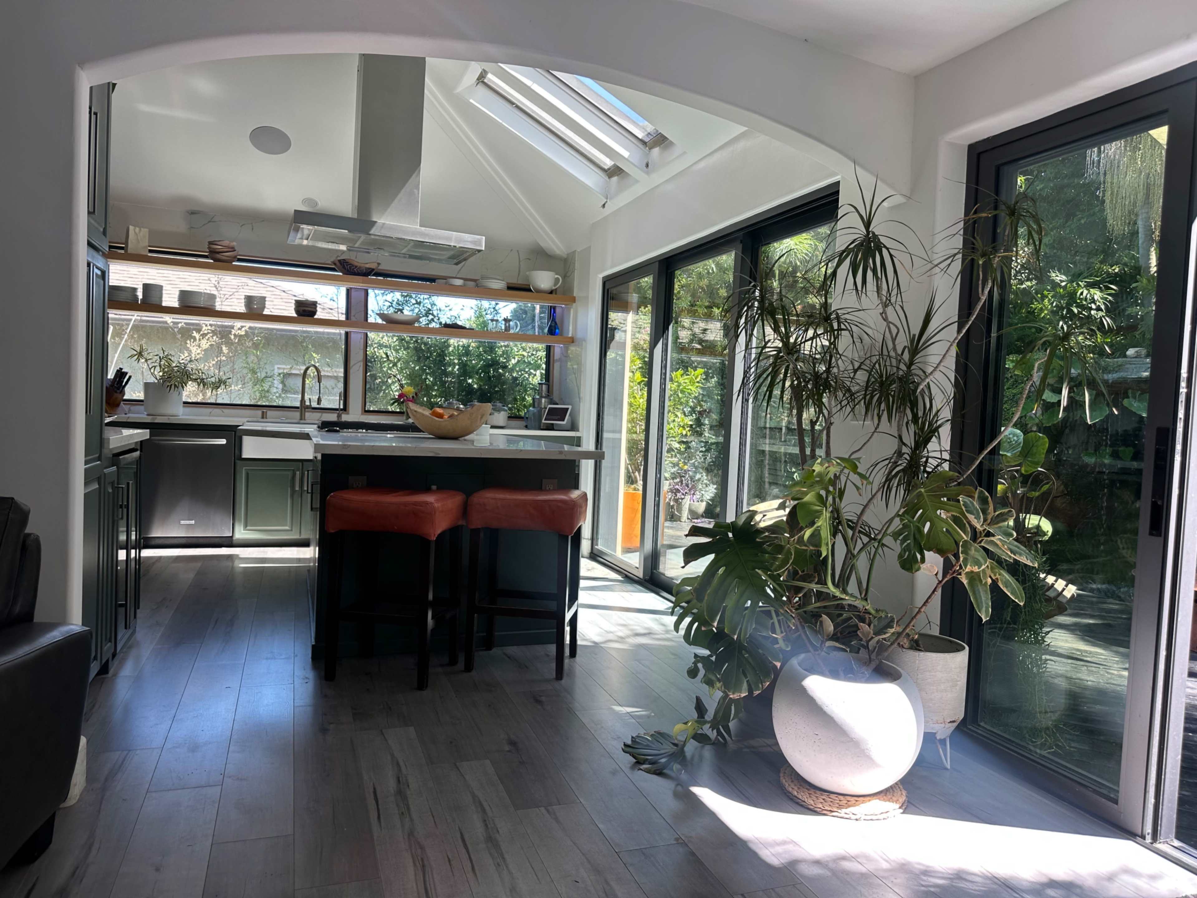 A bright kitchen with a modern design features a large island with bar seating, surrounded by large windows that offer views of greenery outside.