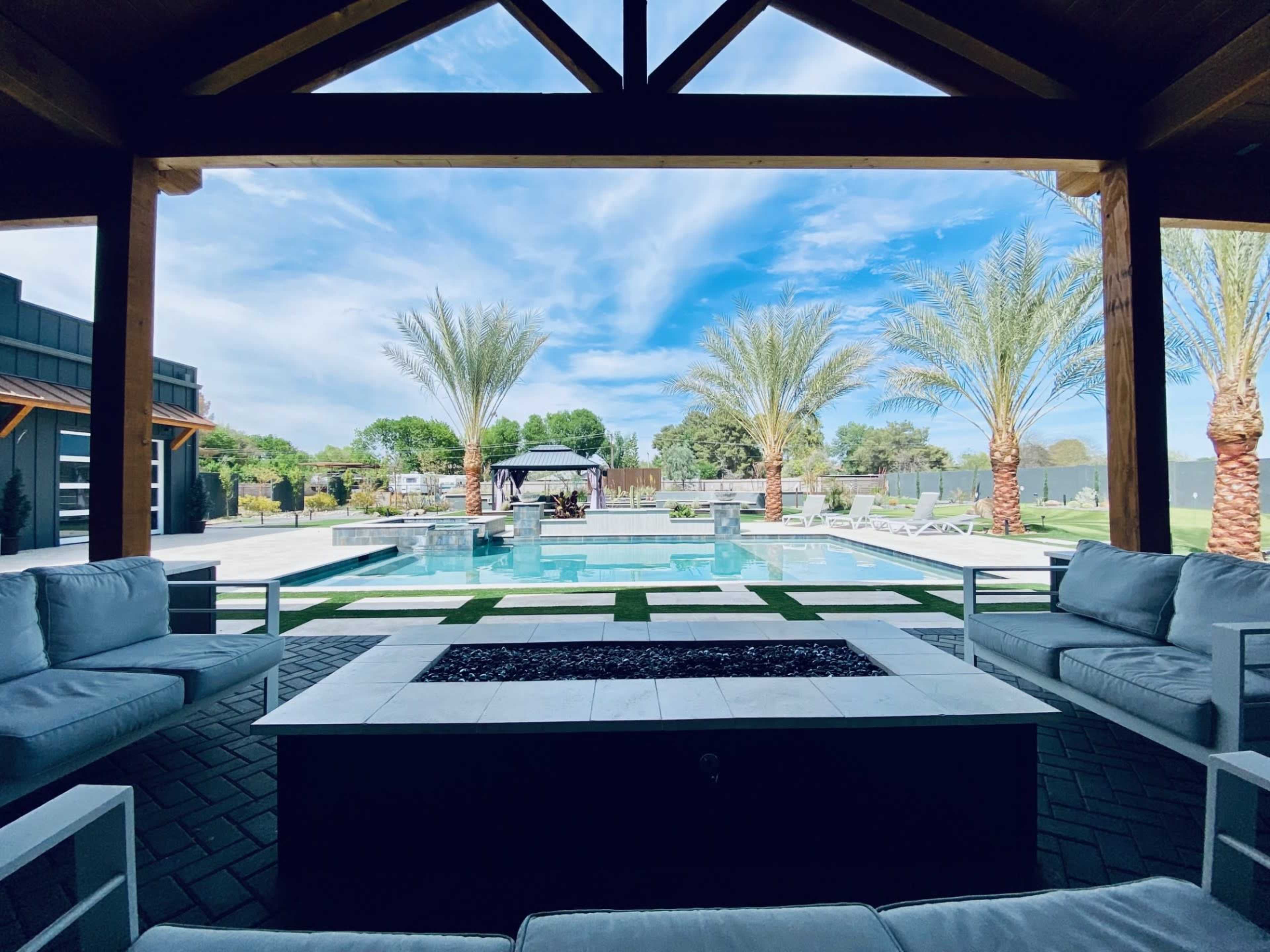 A modern outdoor living space features a pool surrounded by palm trees, patio furniture, and a fire pit under a wooden pergola.