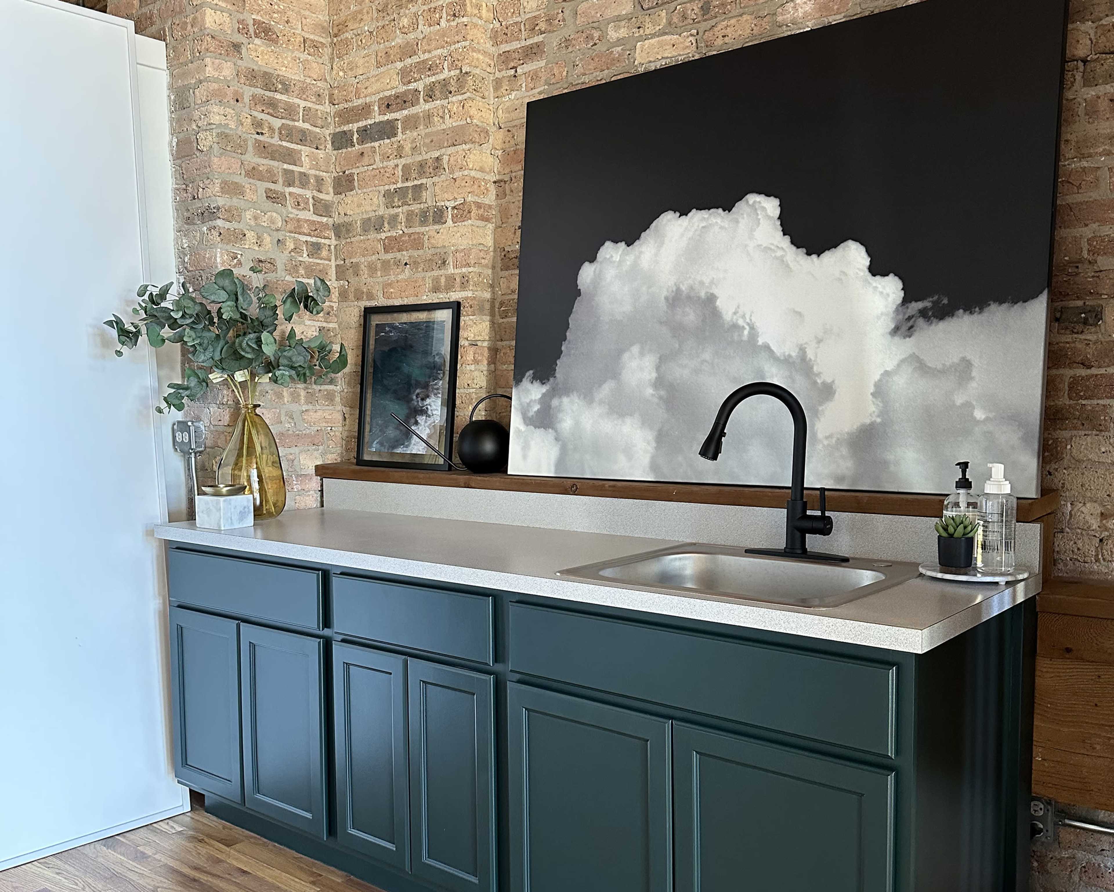 A kitchen countertop features a black faucet, a decorative plant, and a large black-and-white cloud artwork against a brick wall.