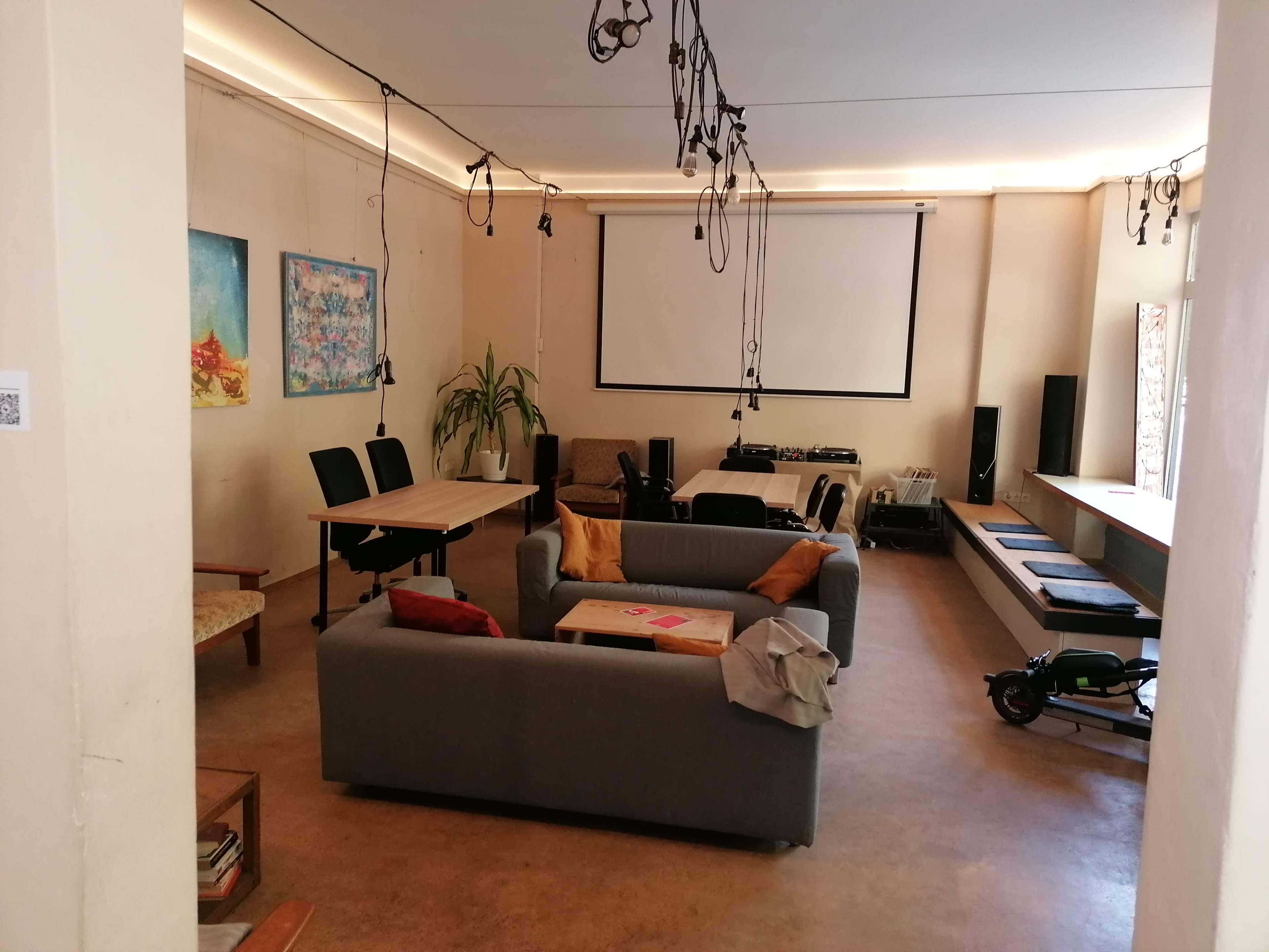 The image shows a spacious room with a gray sofa, a wooden table with chairs, and a projector screen, along with various audio equipment and decorative wall art.