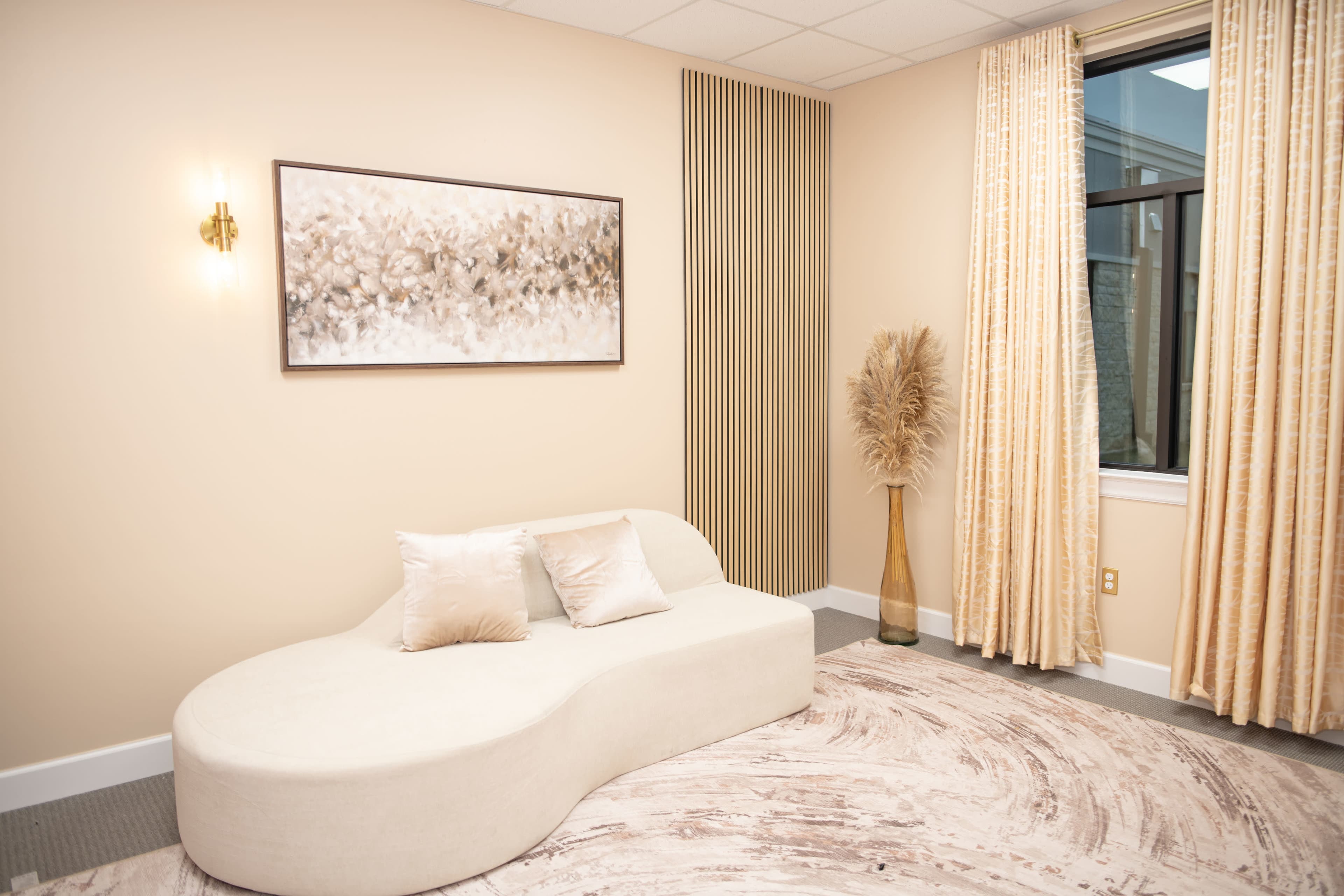 The image shows a softly curved cream-colored sofa positioned against a light beige wall adorned with a floral artwork and complemented by curtains and a decorative vase.