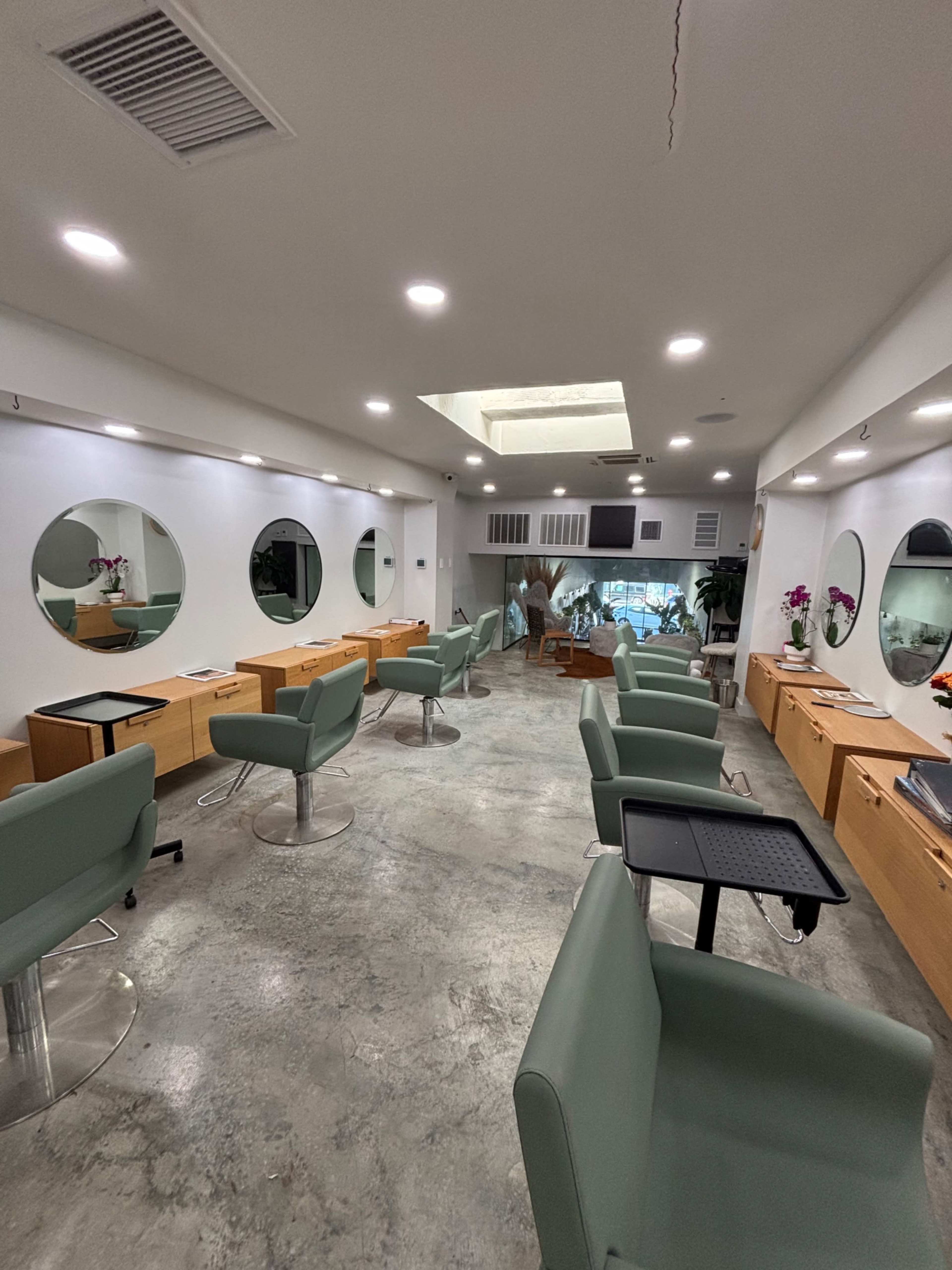 The image shows a modern hair salon with several styling stations, each featuring green chairs and circular mirrors, along with a skylight and decorative plants.