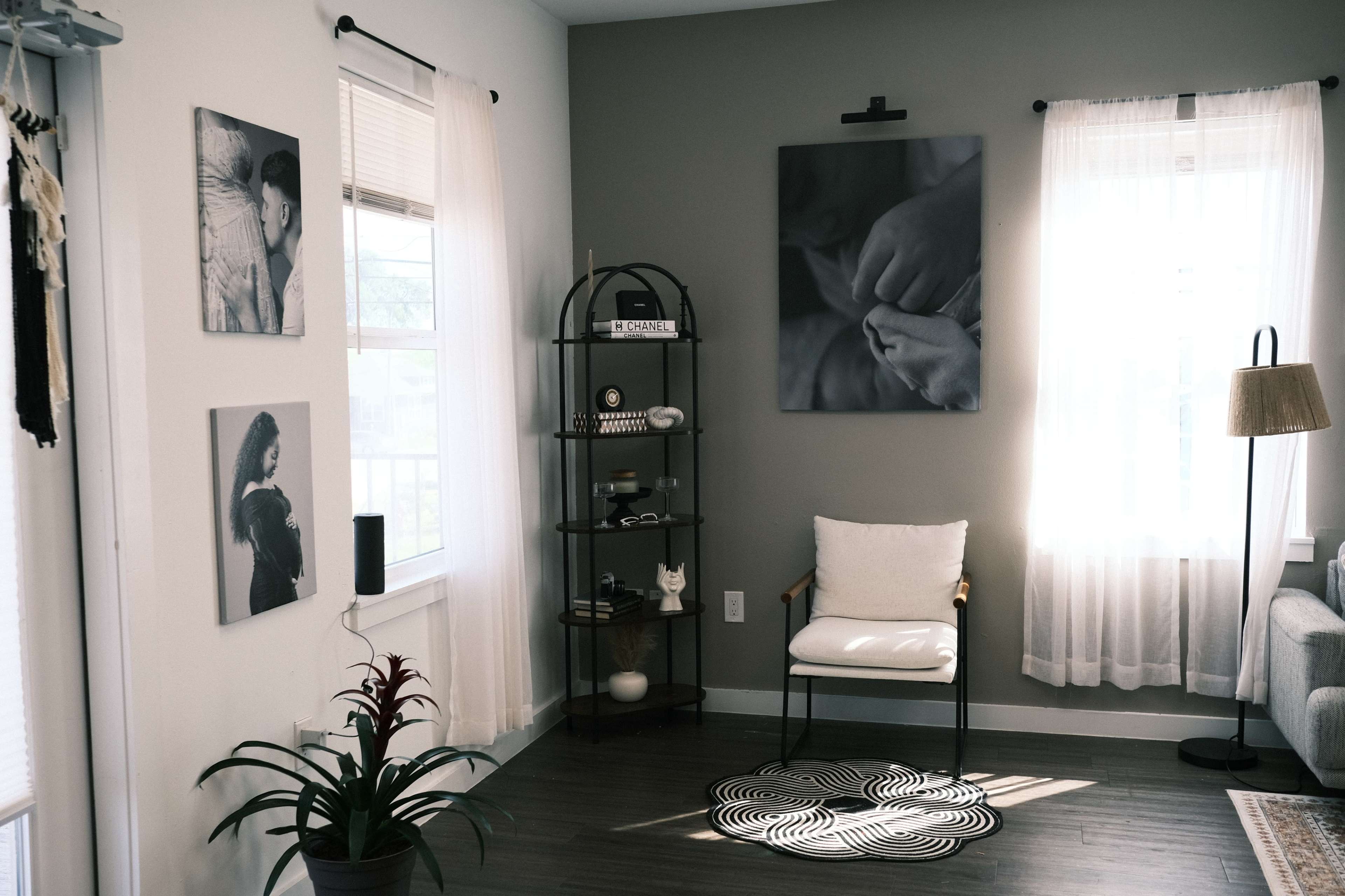 The image depicts a corner of a modern living room featuring a chair, a circular patterned rug, a glass display shelf with decorative items, and framed artwork on the walls.
