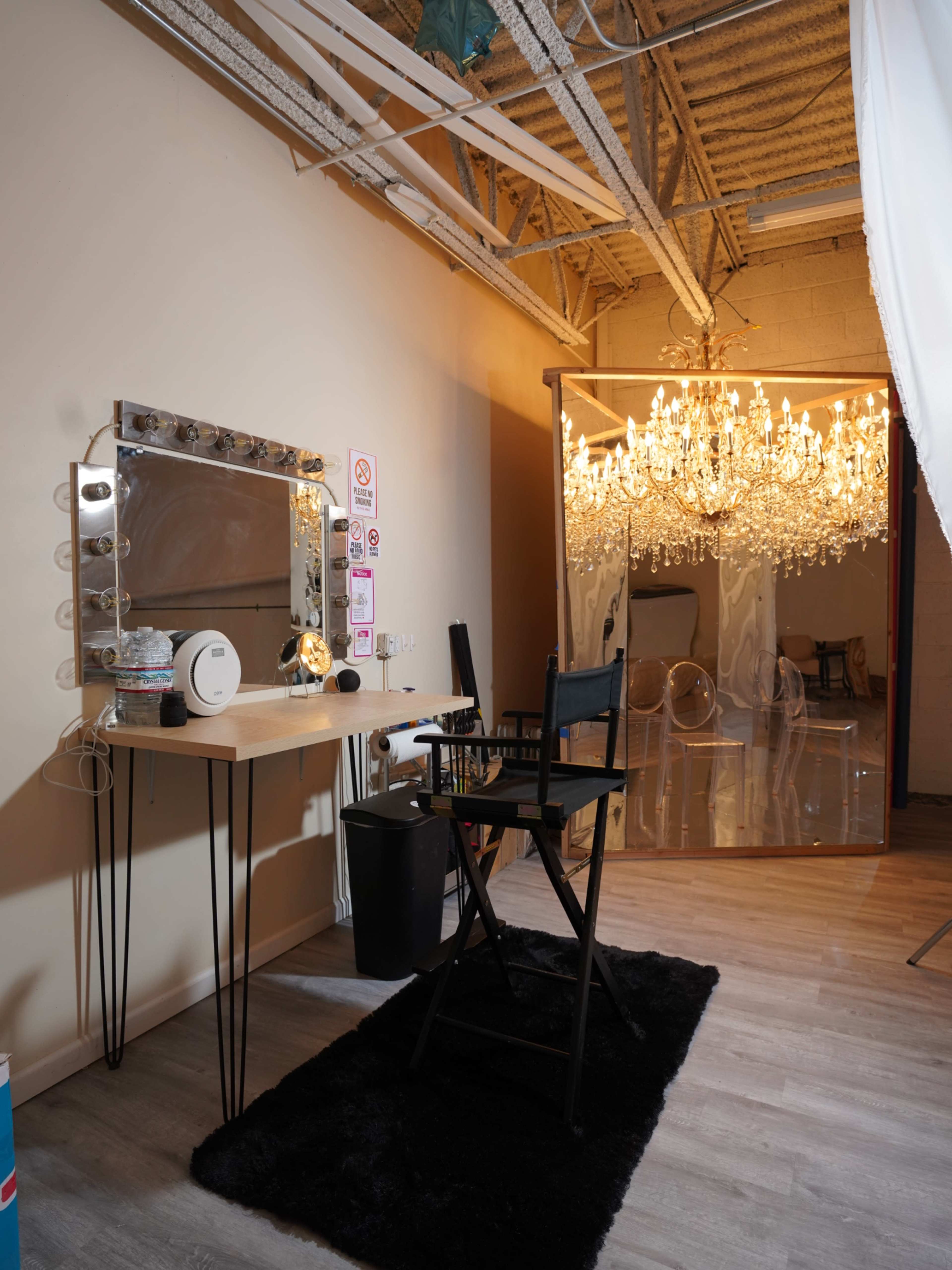 A beauty station with a lit mirror and chair is set against a backdrop featuring a large chandelier.