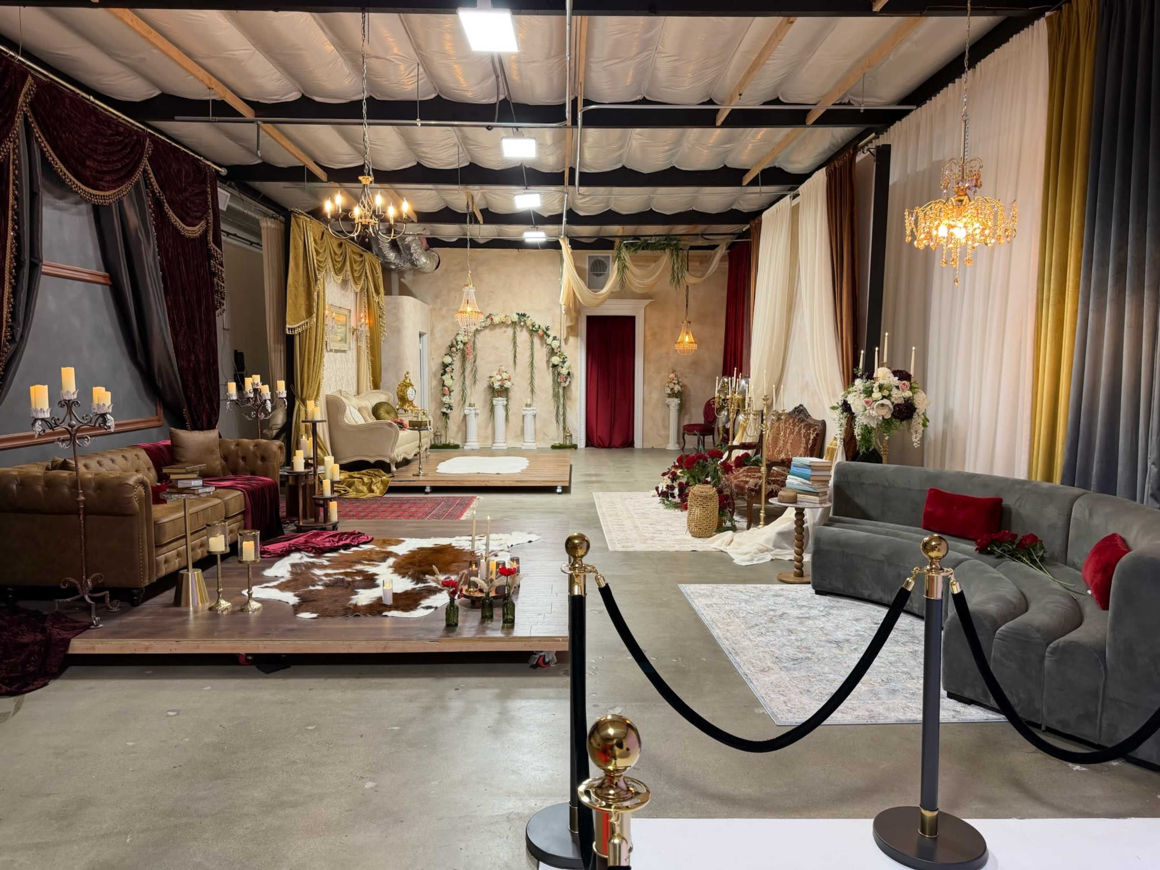 The image shows a spacious event venue decorated with various lounge areas featuring elegant furniture, floral arrangements, and dramatic drapery.