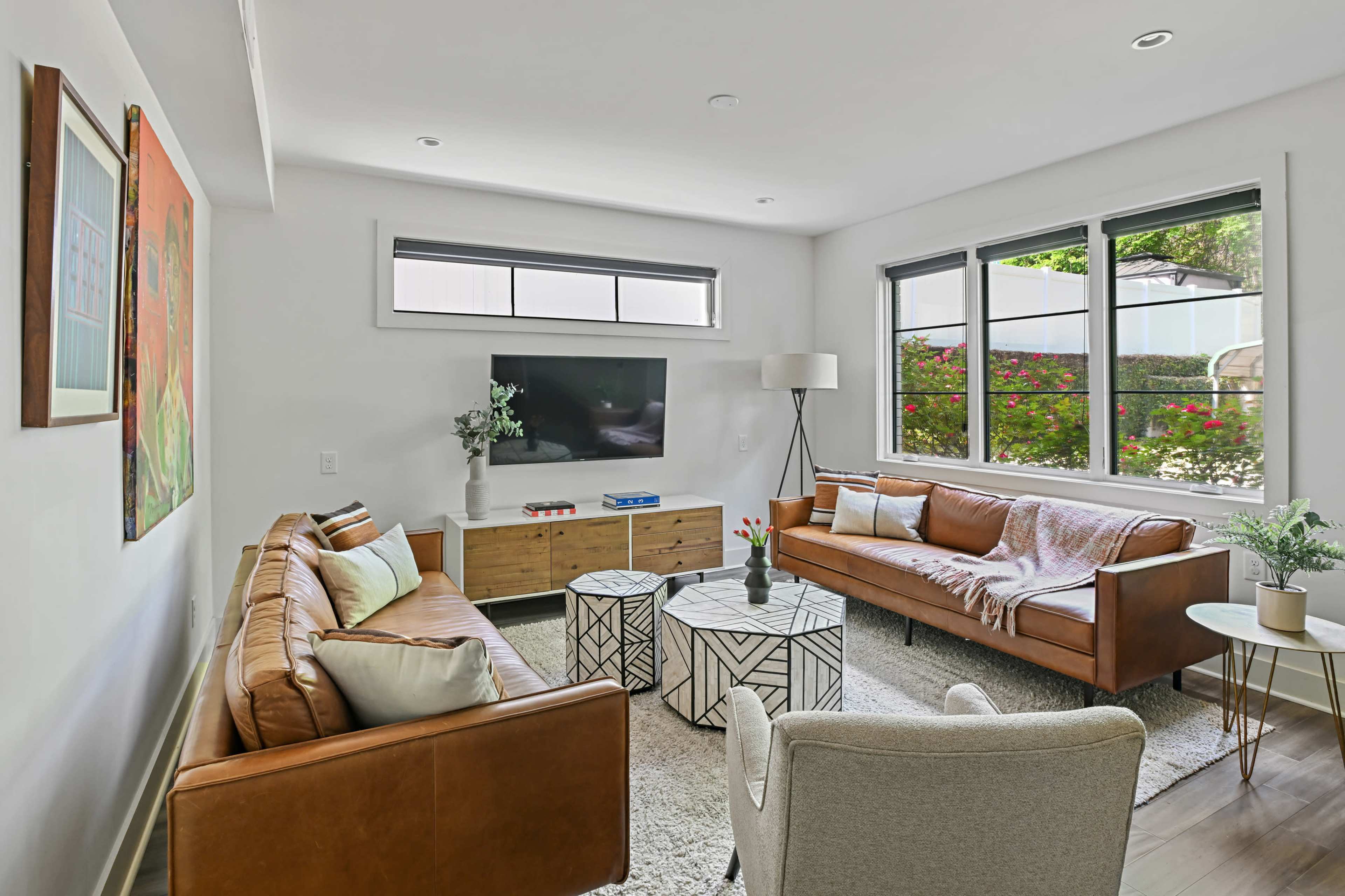 The image shows a modern living room with two leather sofas, a coffee table, a wall-mounted television, and large windows overlooking a garden.