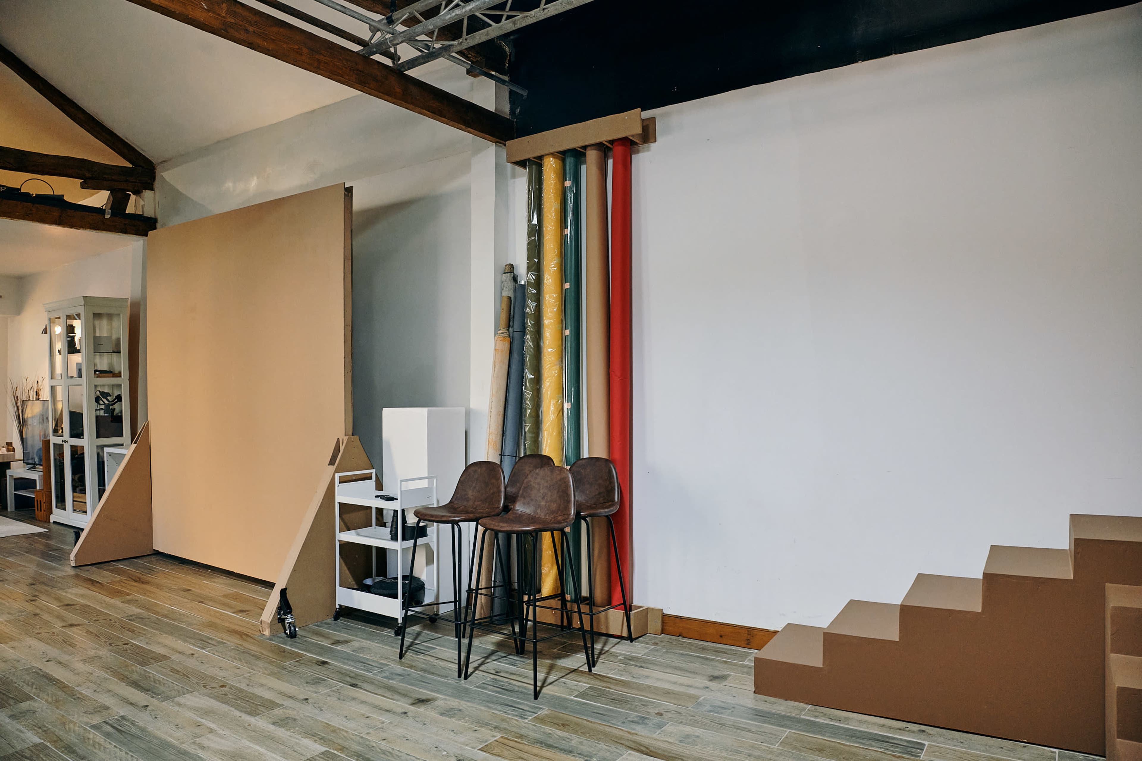 The image shows a studio space with a large beige backdrop, colorful rolls of paper, a set of three bar stools, and a minimalist staircase made of cardboard.