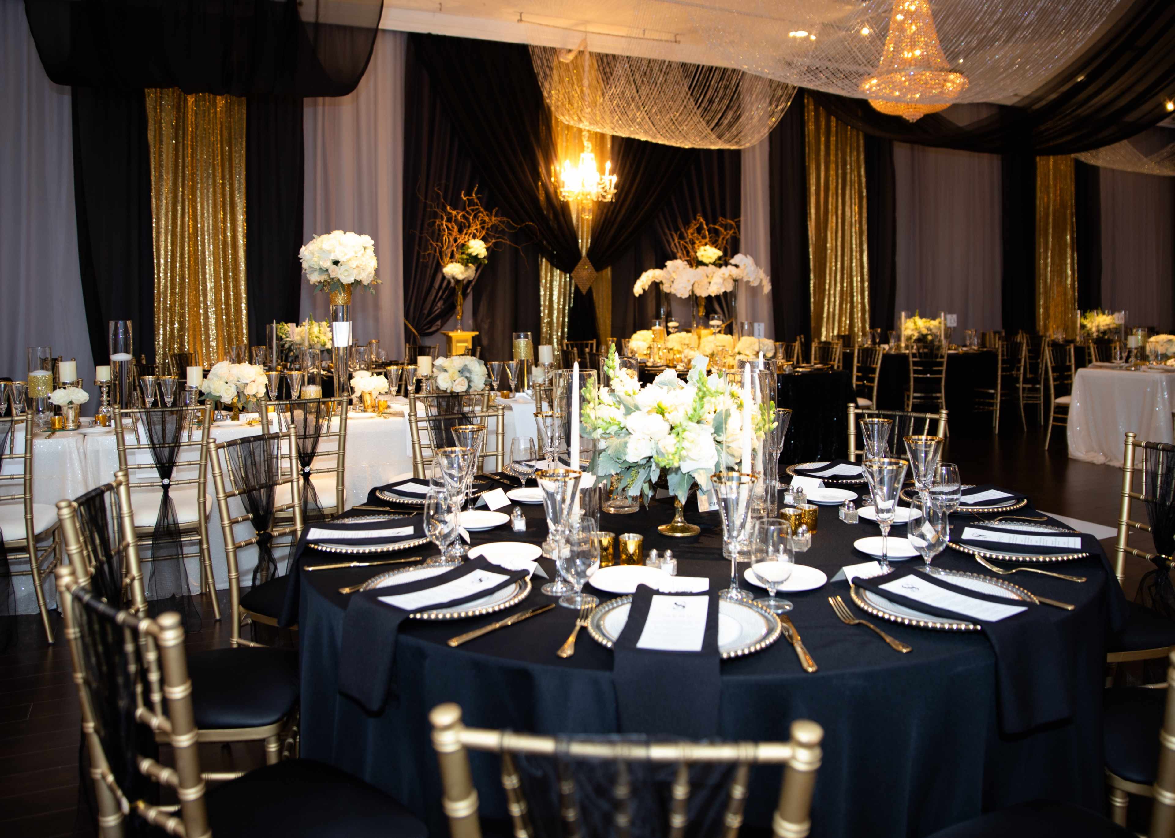 A formal dining setup is arranged in a banquet hall, featuring tables adorned with black tablecloths, white floral centerpieces, and golden accents against a backdrop of draped fabric.