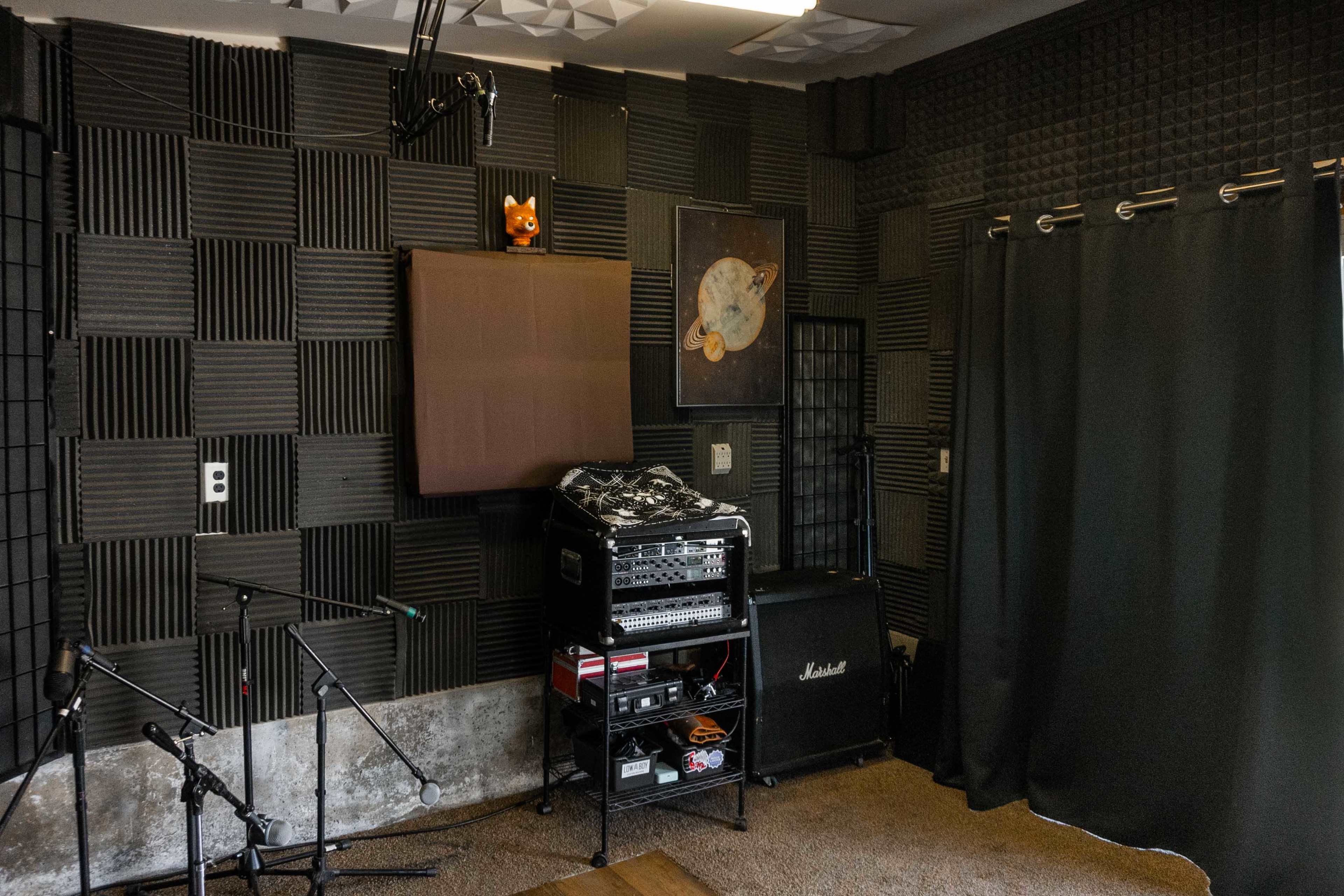 A soundproofed recording studio with black acoustic wall panels, a rack of audio equipment, a Marshall amplifier, and a dark curtain.