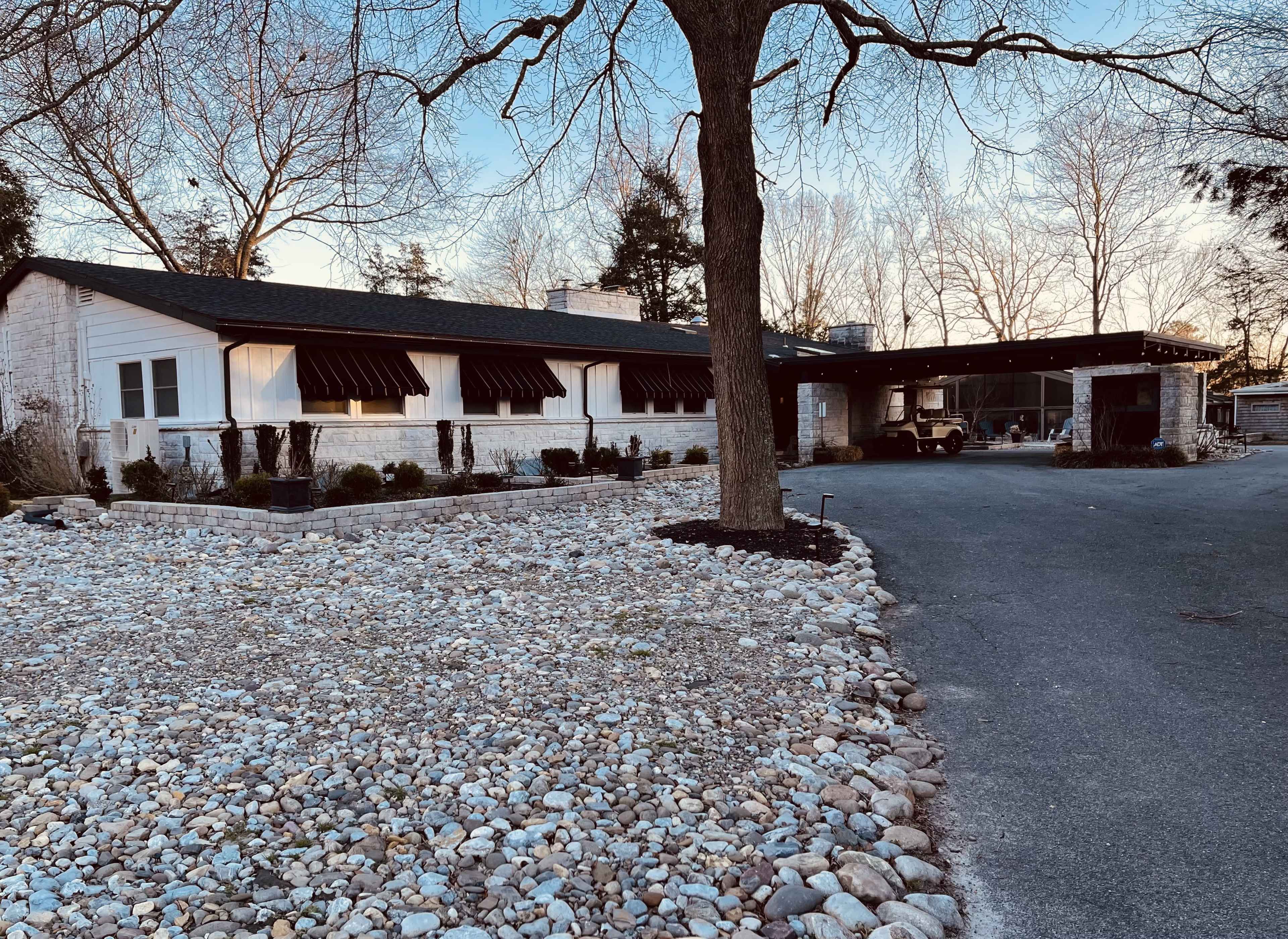 A single-story house with a stone exterior and a covered porch is situated beside a gravel driveway surrounded by trees and landscaping.