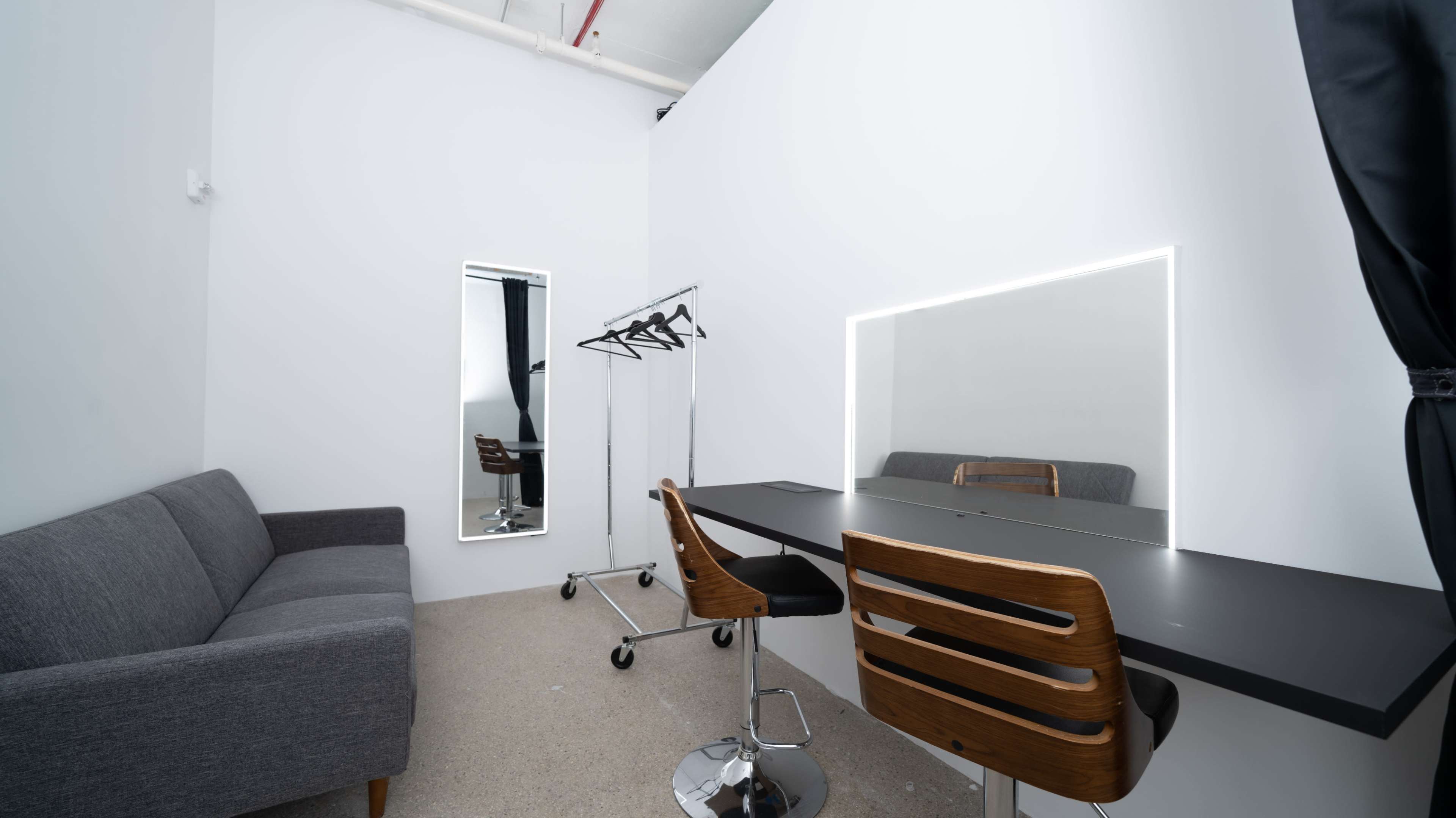 A minimalistic room featuring a gray sofa, a mirror, a clothing rack, and a desk with two wooden chairs.