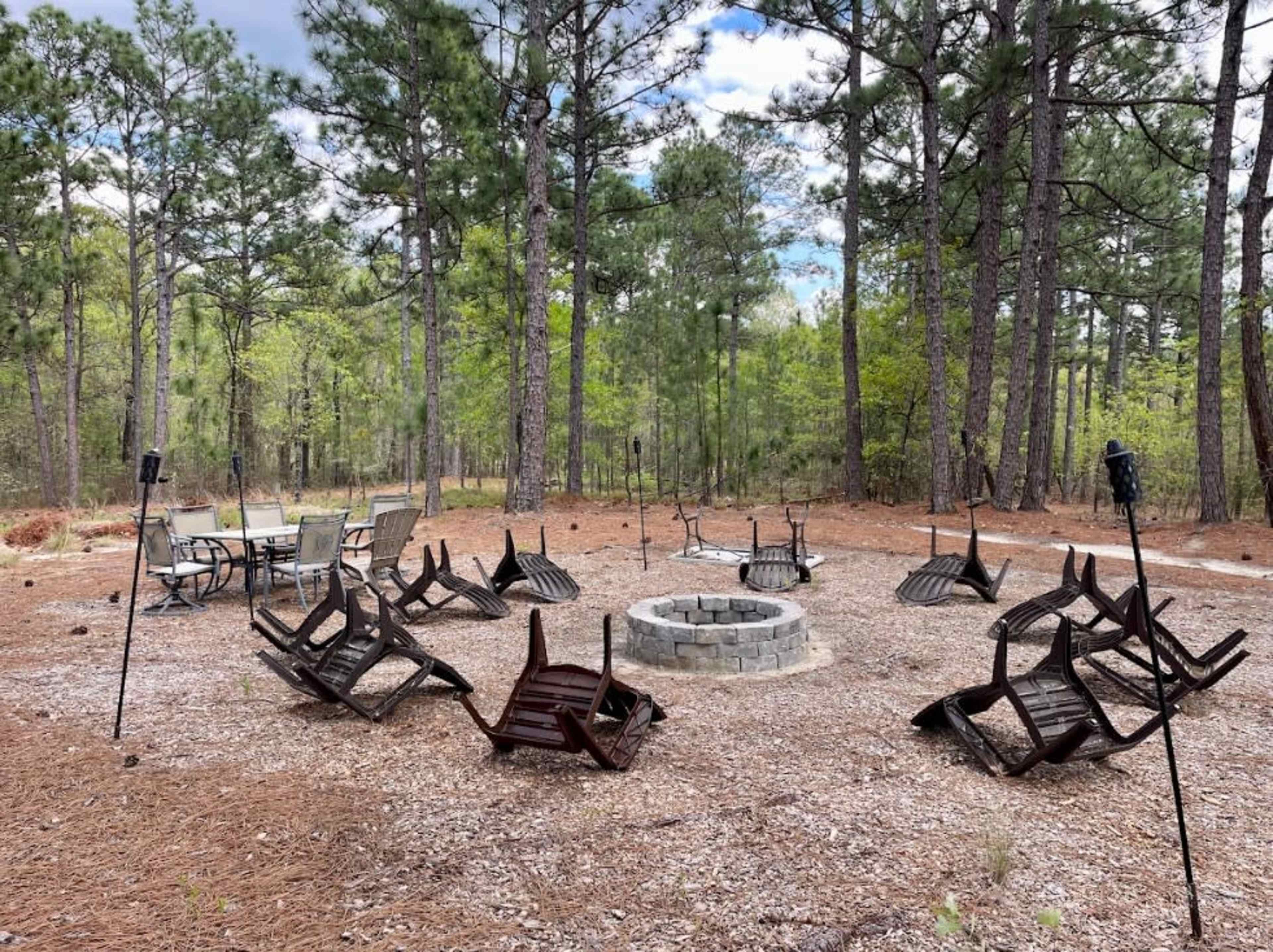 A circular outdoor fire pit area is surrounded by overturned wooden chairs and a table, set among tall pine trees.