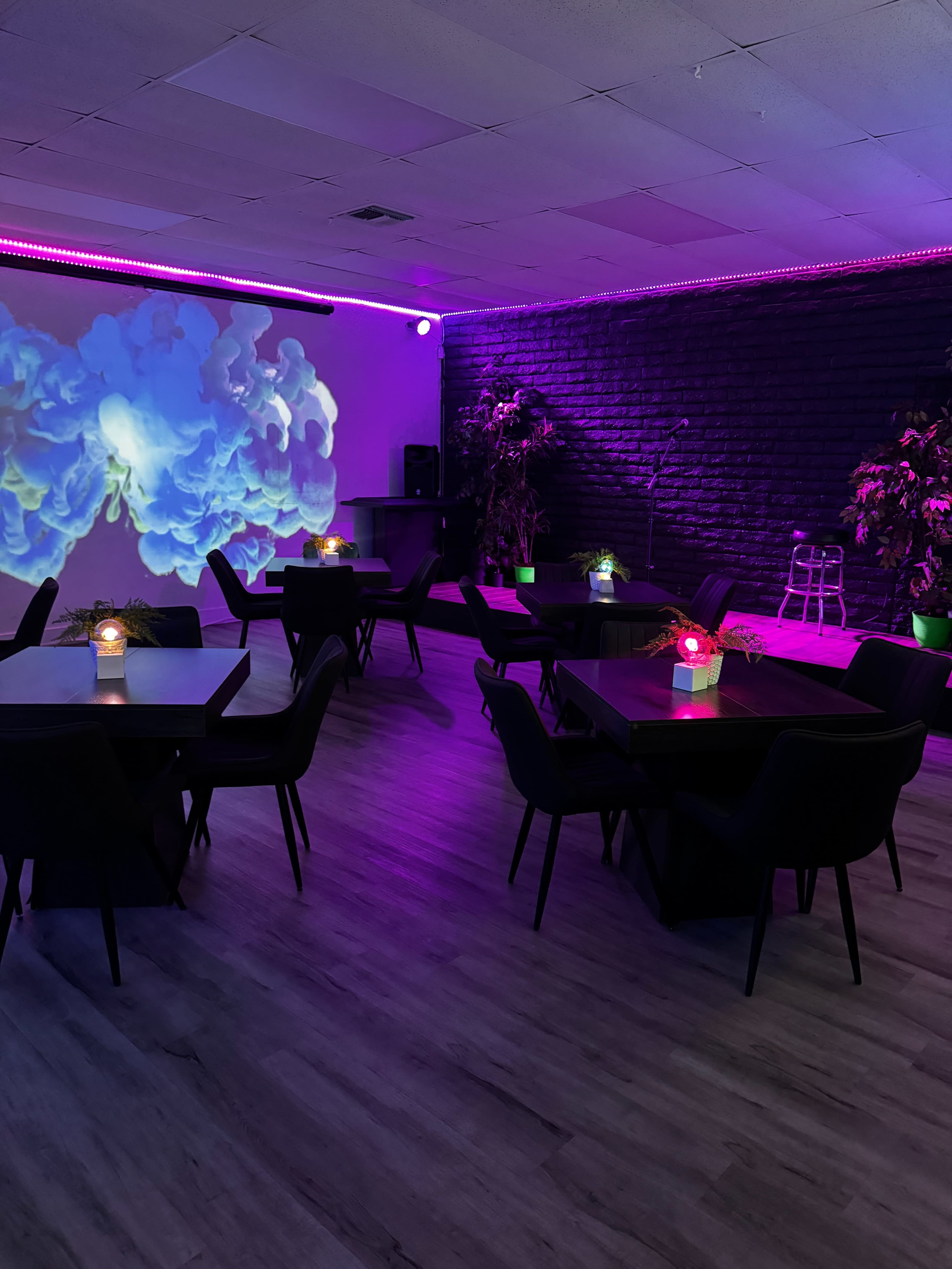 The image shows a dimly lit room with black tables and chairs, colorful ambient lighting, and a projection of swirling clouds on the wall.