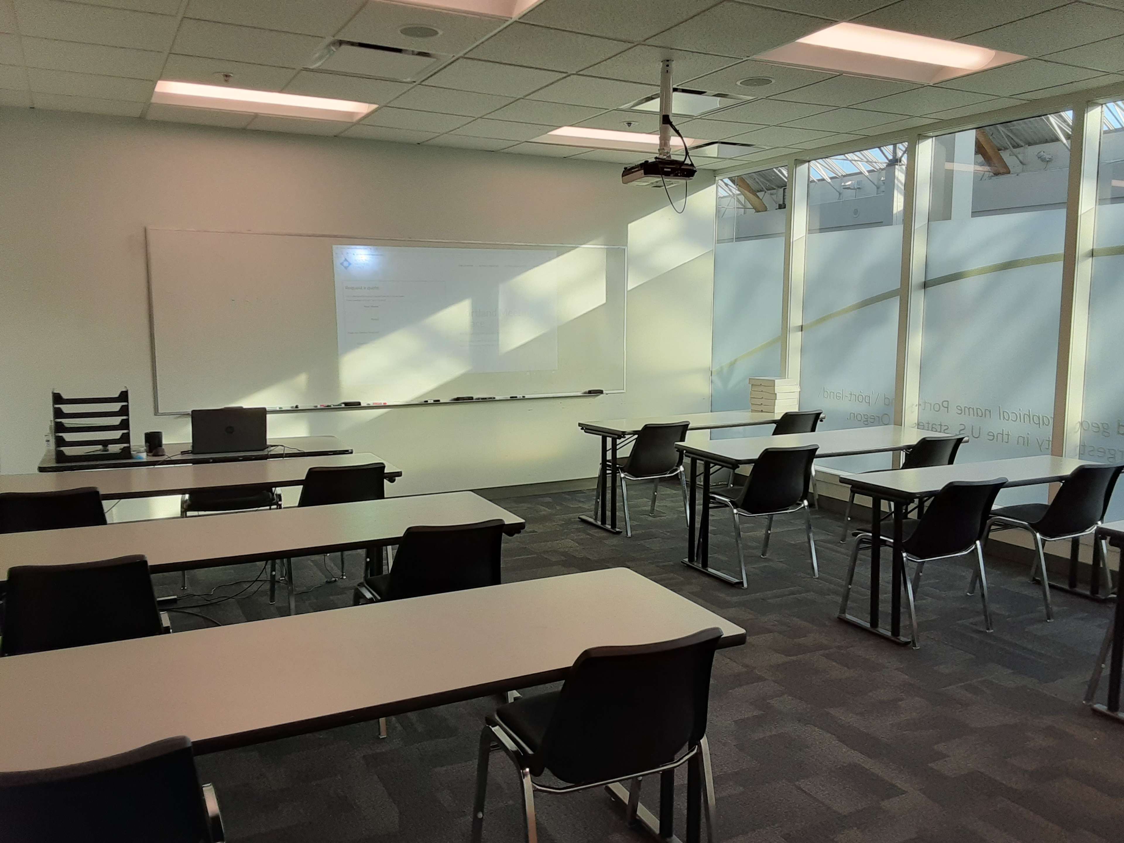 A classroom setup with rows of empty desks, a projection screen, and large windows allowing natural light.