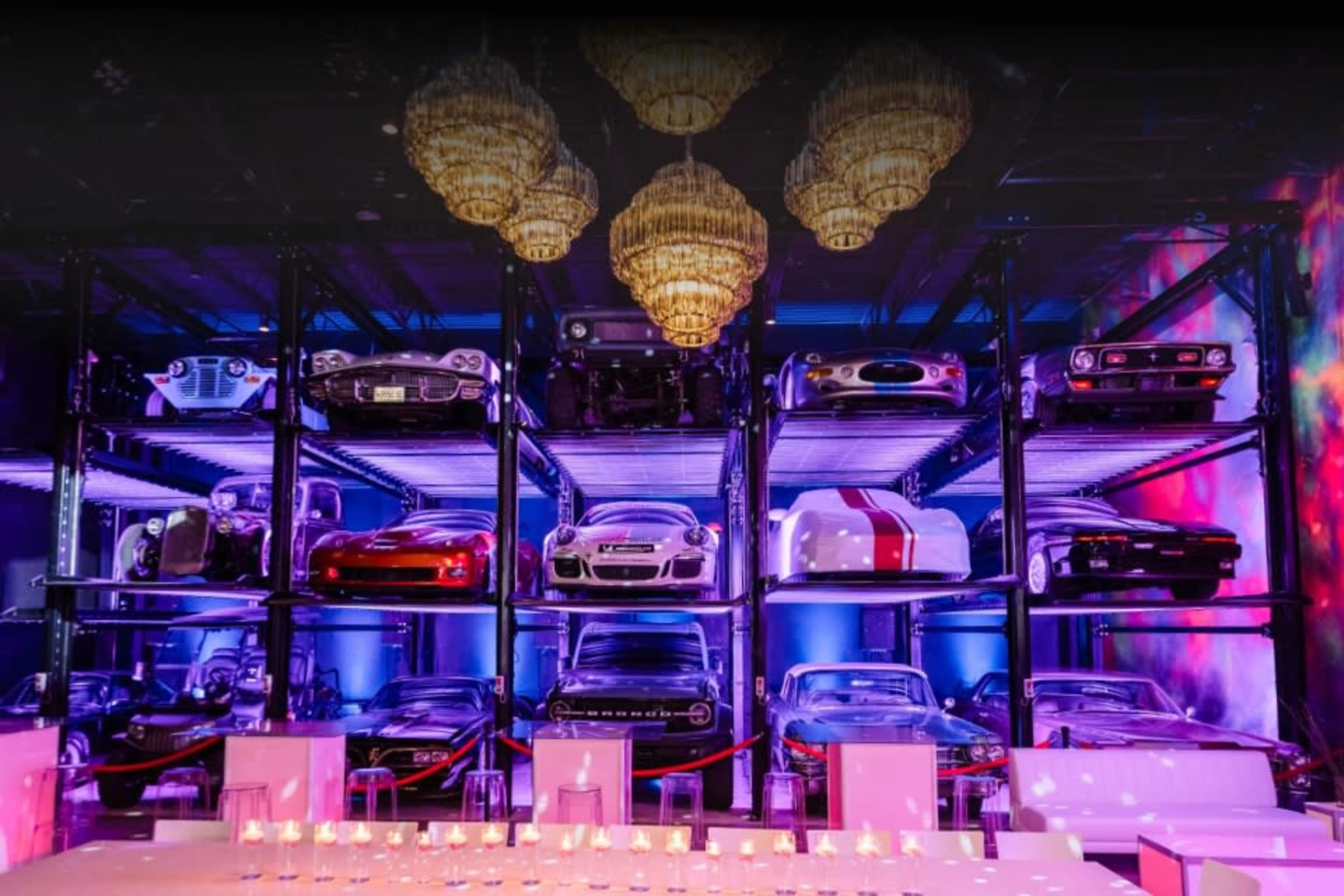 A display of various classic and modern cars is arranged on shelves under illuminated chandeliers in a stylish event space.