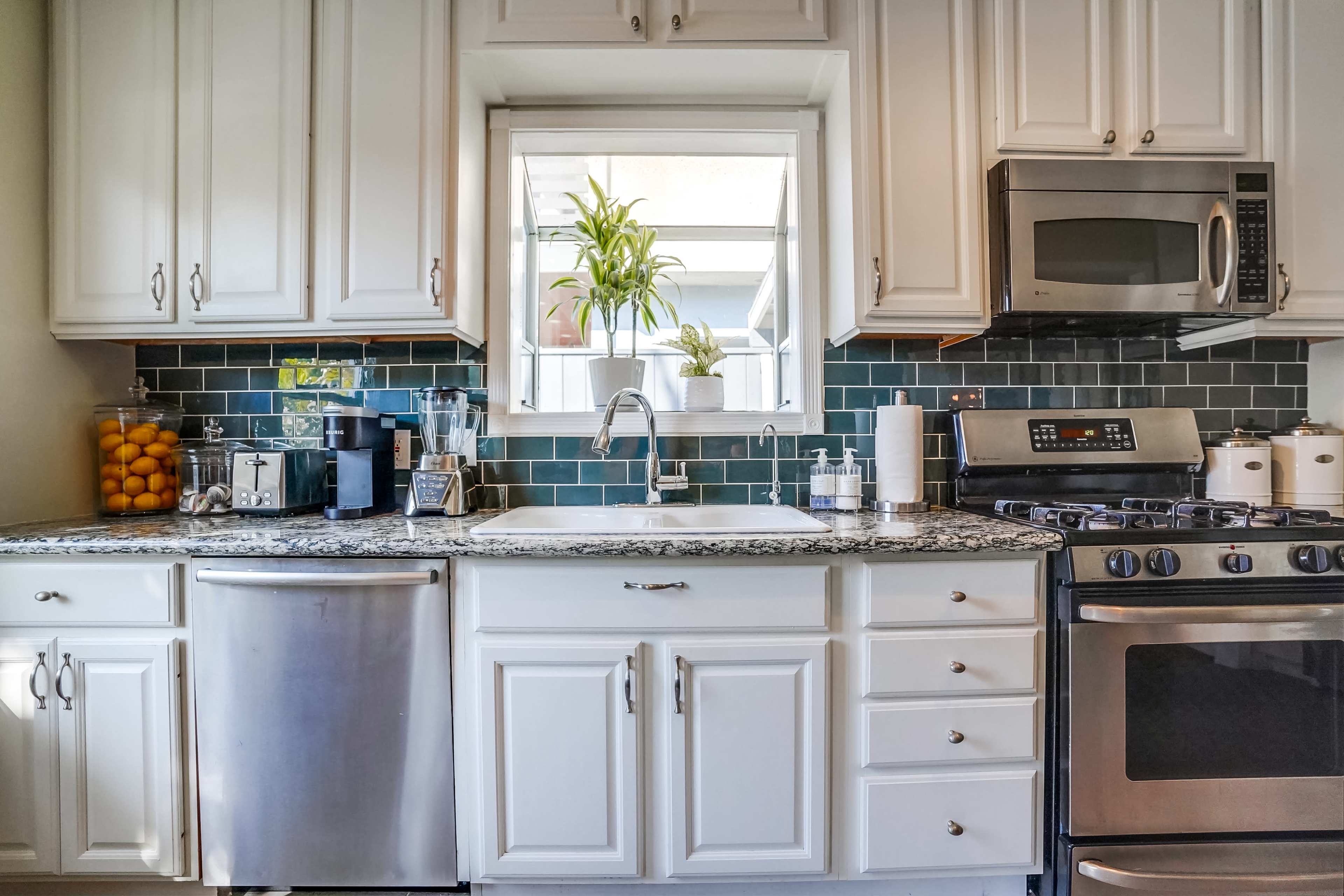 A modern kitchen features white cabinets, a stainless steel sink, and appliances, with a window displaying a potted plant.