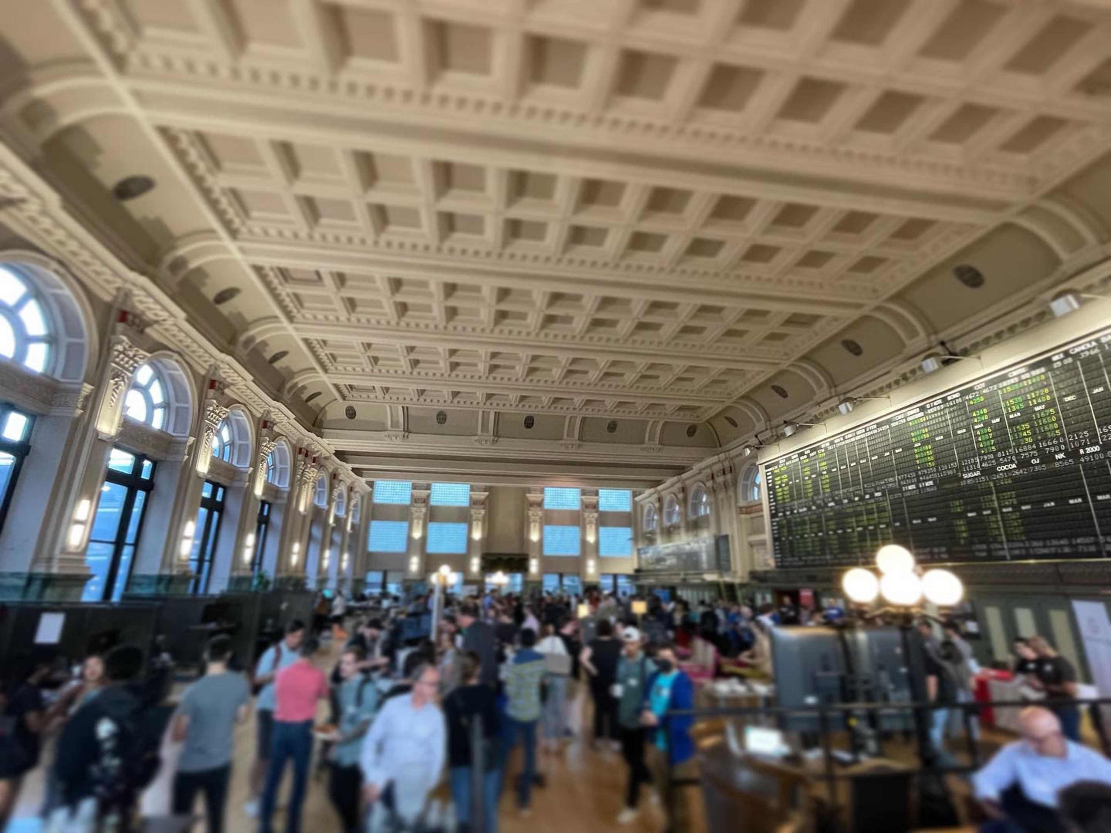 A crowded train station interior features a high, ornate ceiling and large windows, with a digital display board visible at one end.