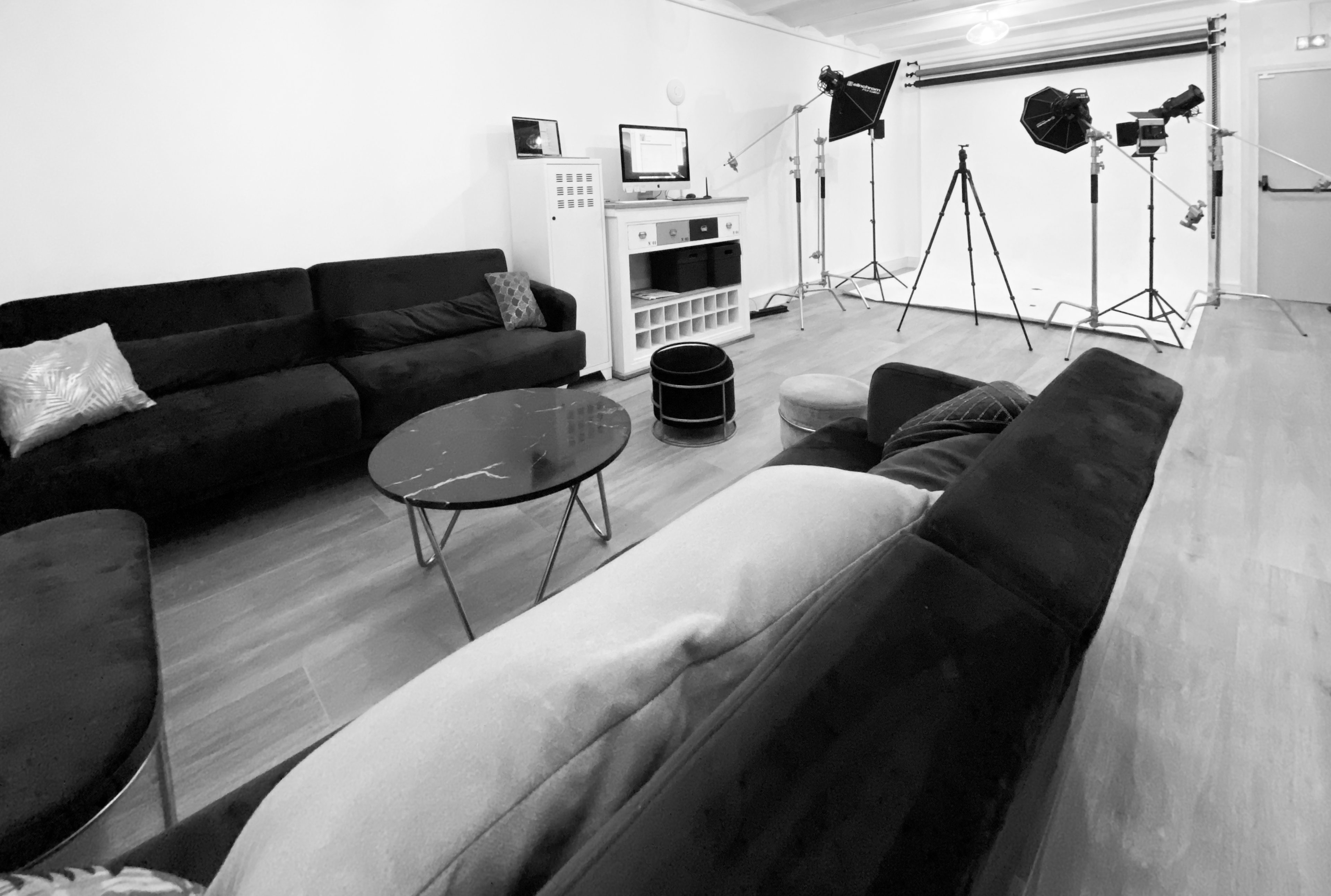 A modern living room setup with black sofas, a coffee table, and studio lighting equipment positioned in the background.