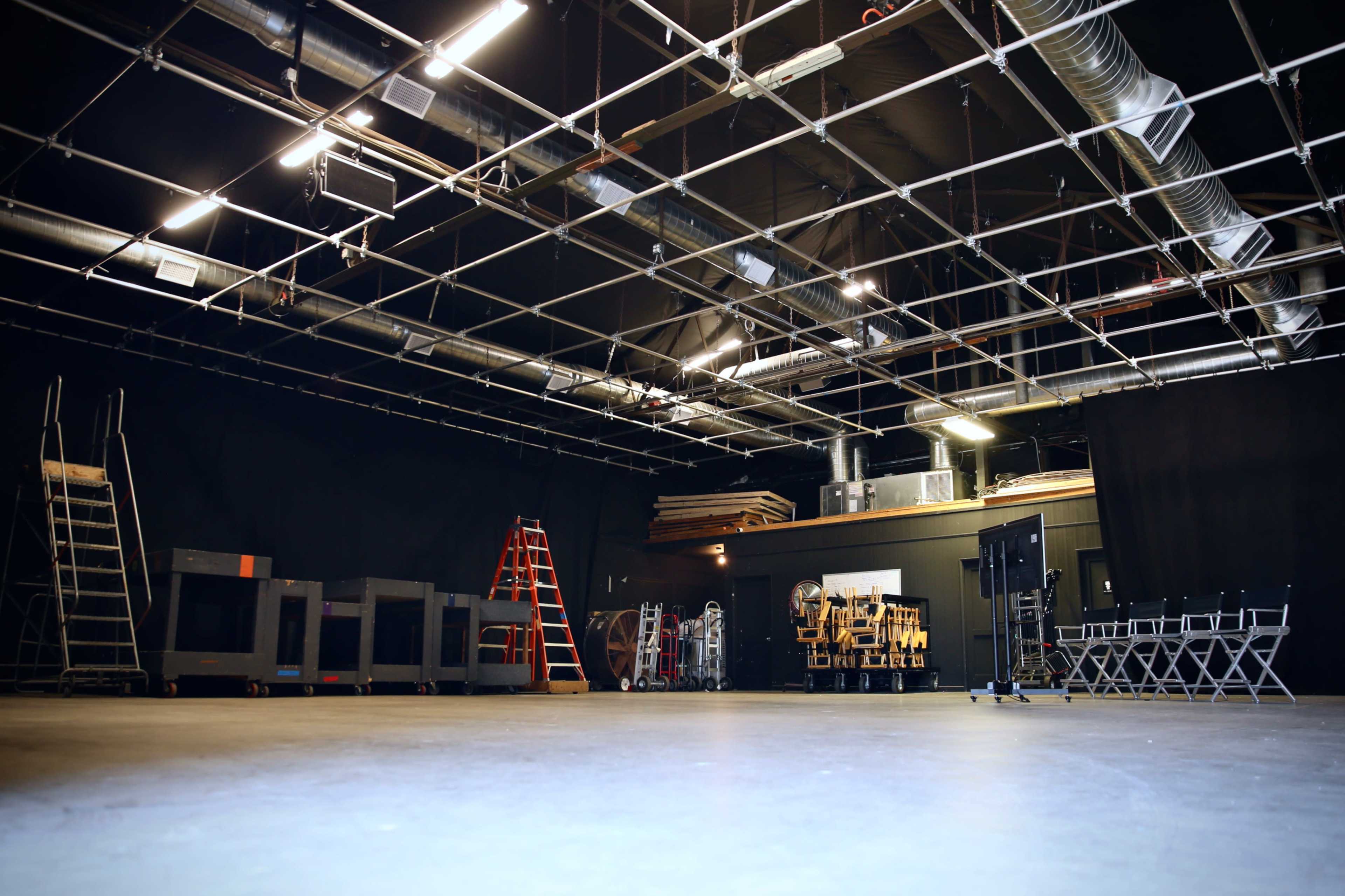 An empty film studio with overhead lighting, black curtains, and various equipment including ladders and carts arranged along the walls.