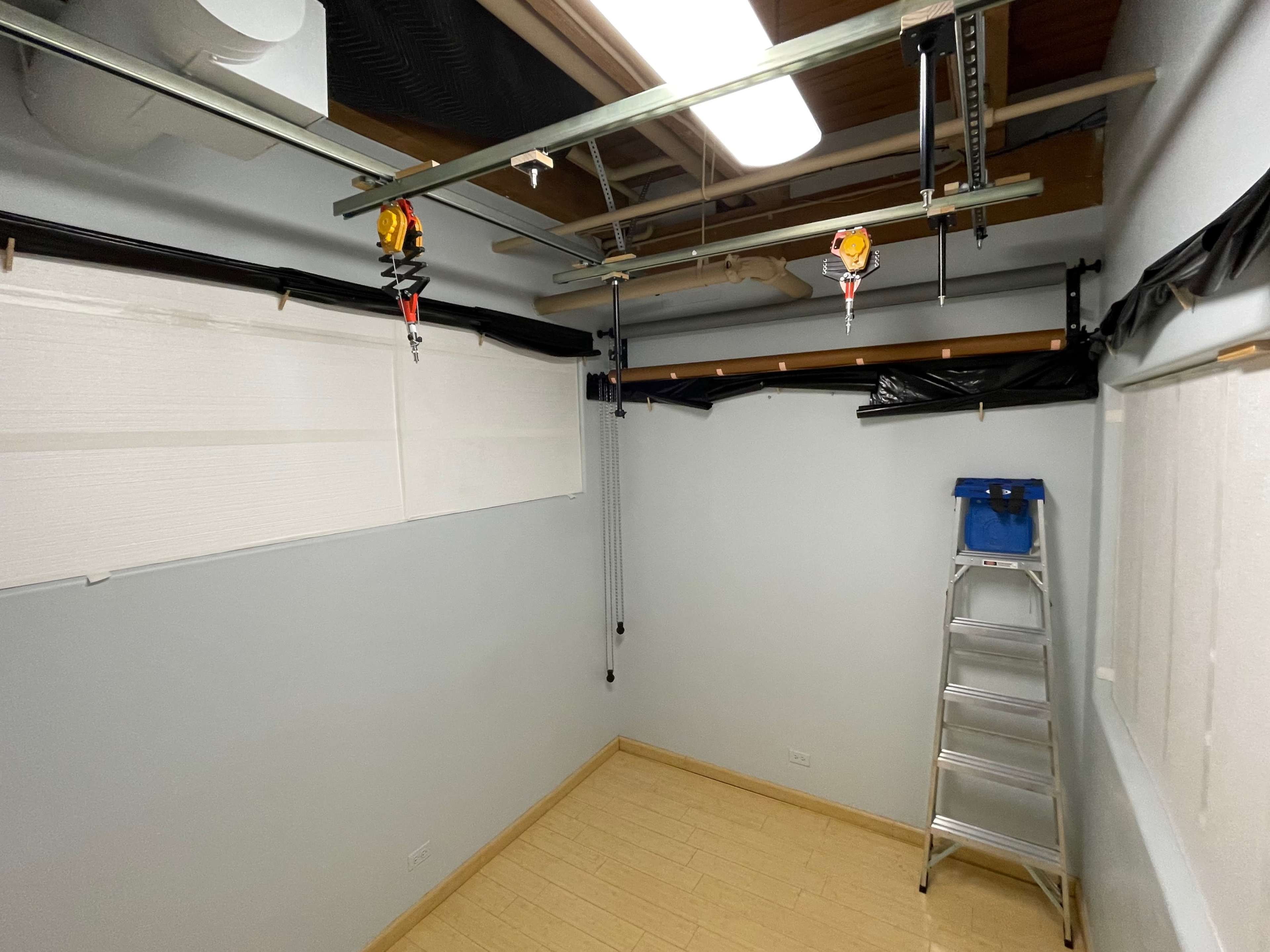 A sparsely furnished room with a ladder, a retractable clothesline, and partially covered windows.