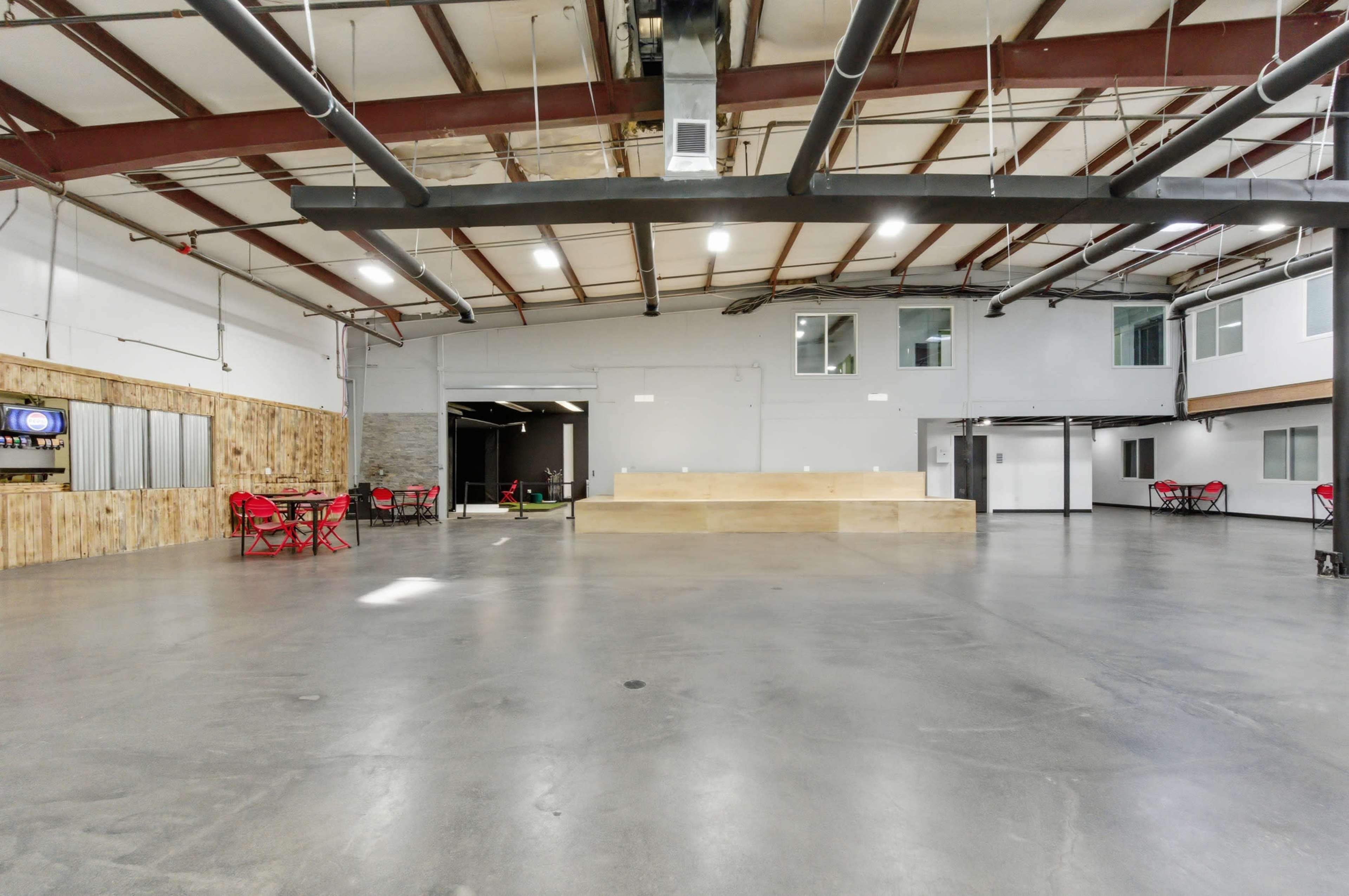 The image shows a spacious industrial-style interior with a concrete floor, exposed steel beams, and areas for seating and activities.