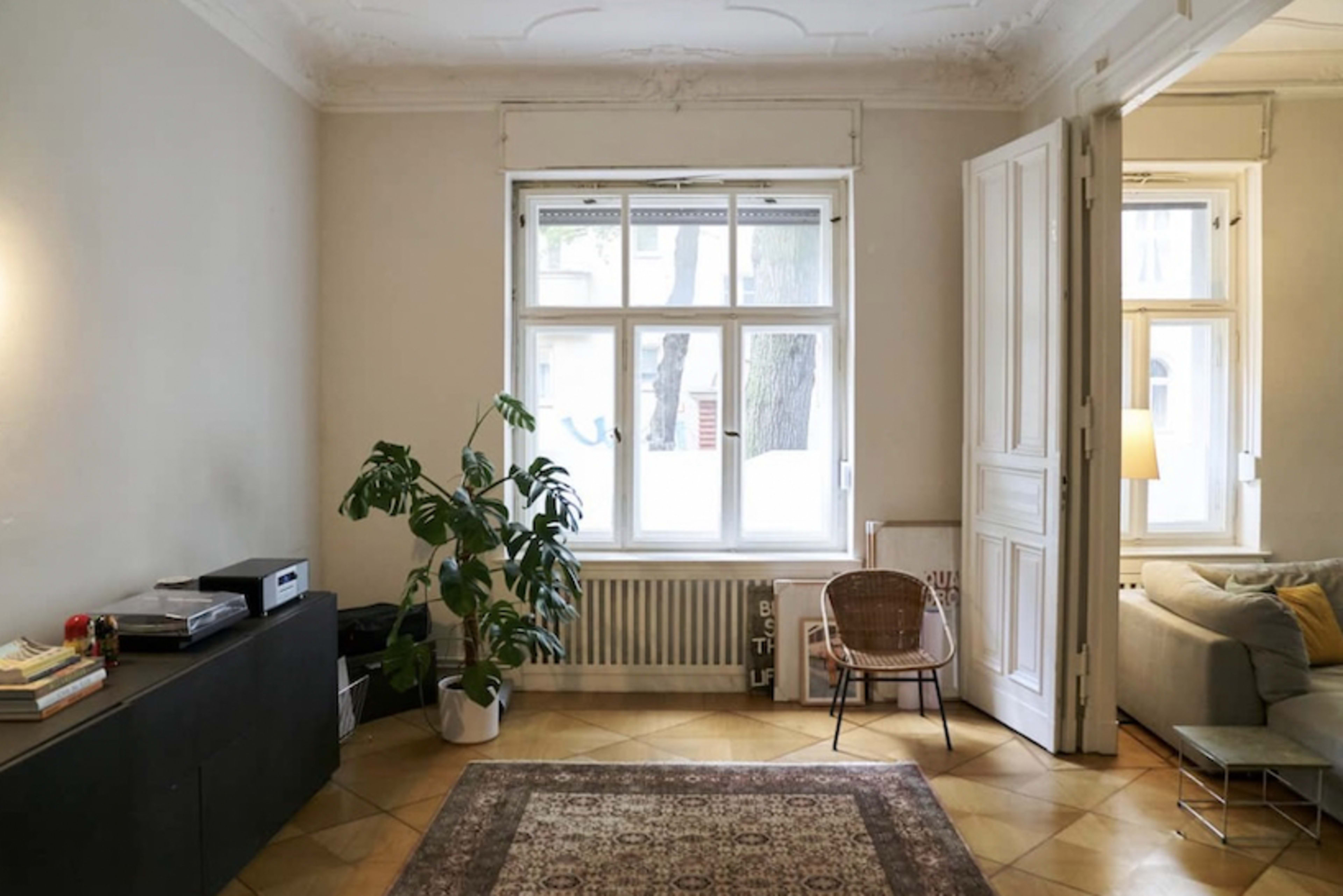 A spacious living room features a large window, a potted plant, a chair, and a sideboard with books.