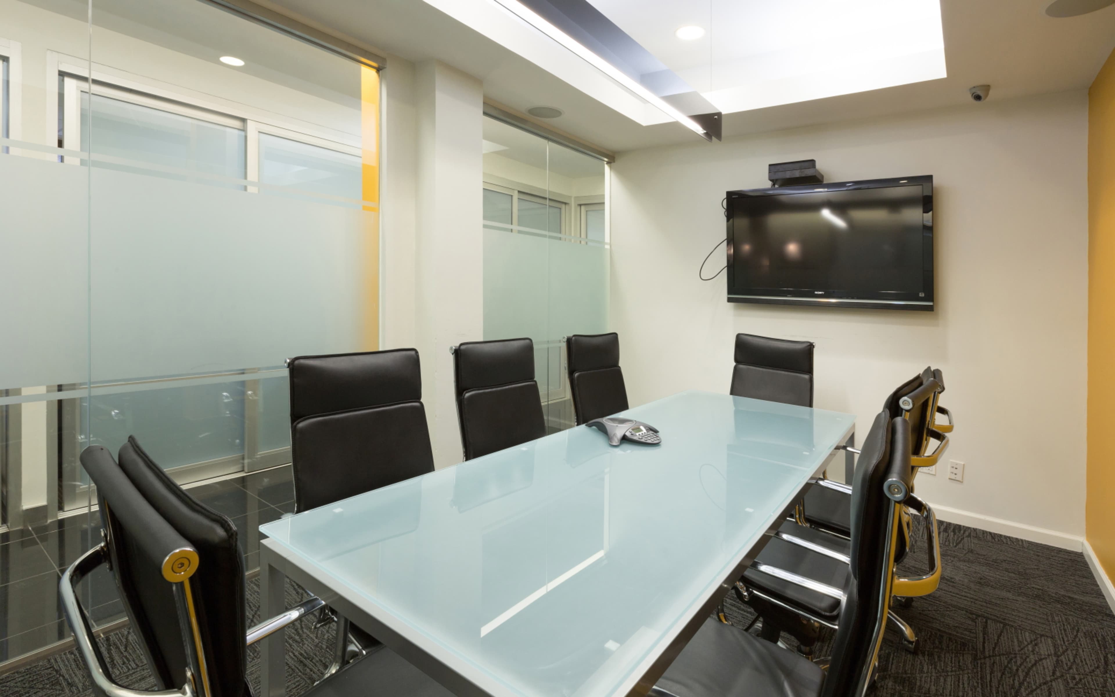 A modern conference room features a glass table surrounded by black chairs and a wall-mounted television.