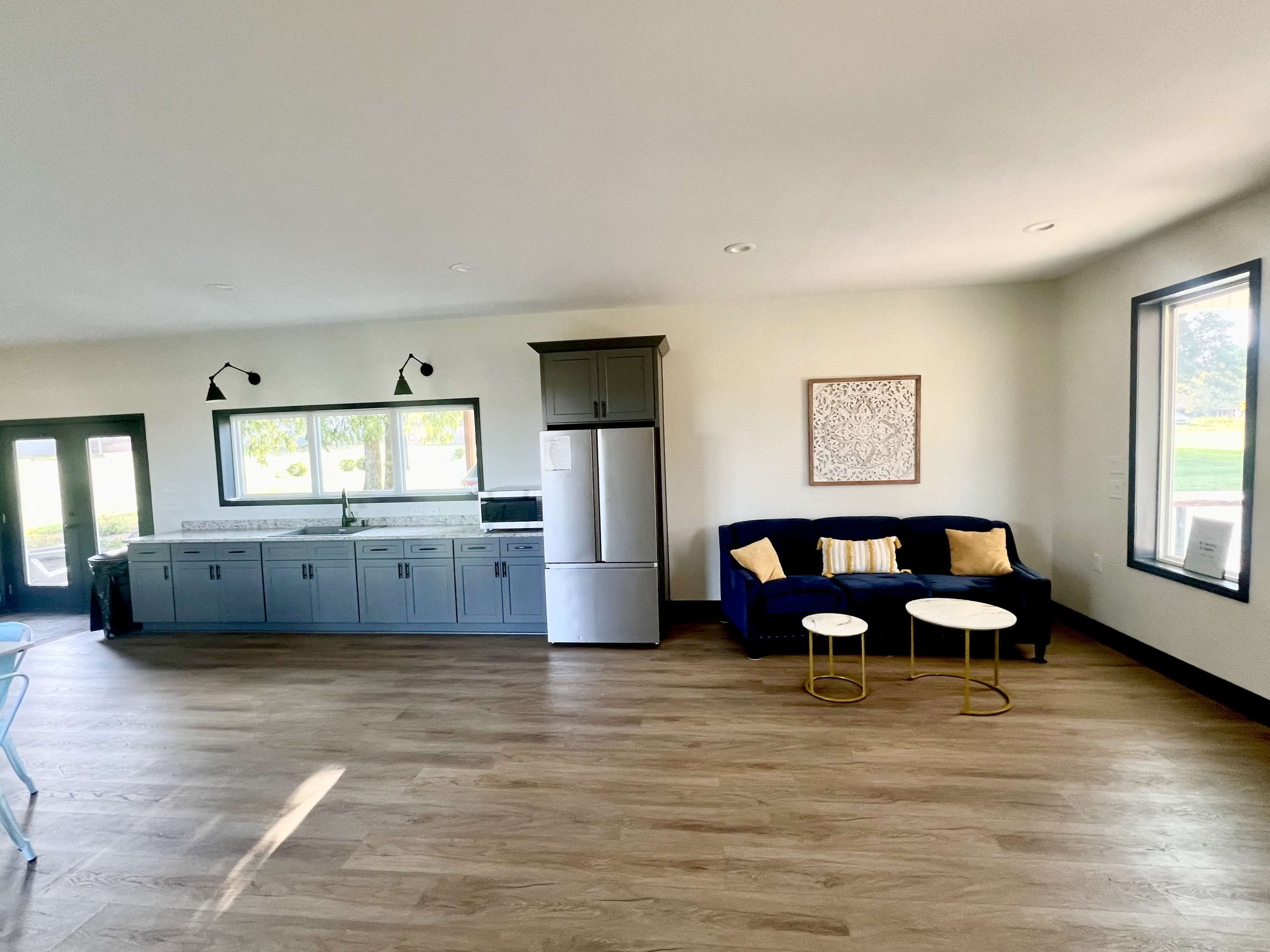 The image shows a modern living space featuring a kitchen area with gray cabinets and a stainless steel refrigerator, along with a blue couch and two round coffee tables.