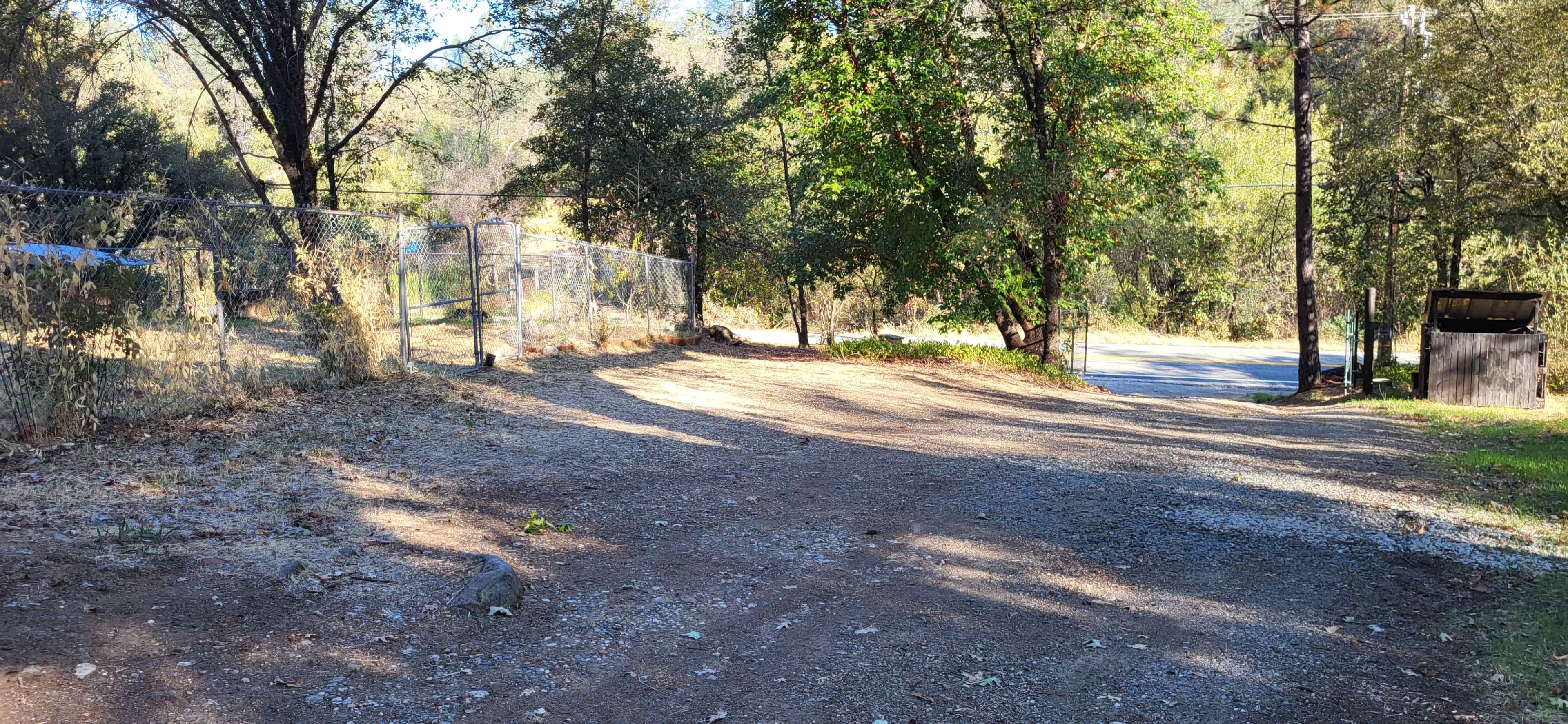 The image shows a gravel path leading towards a fence on the left and a road visible on the right, surrounded by trees and foliage.