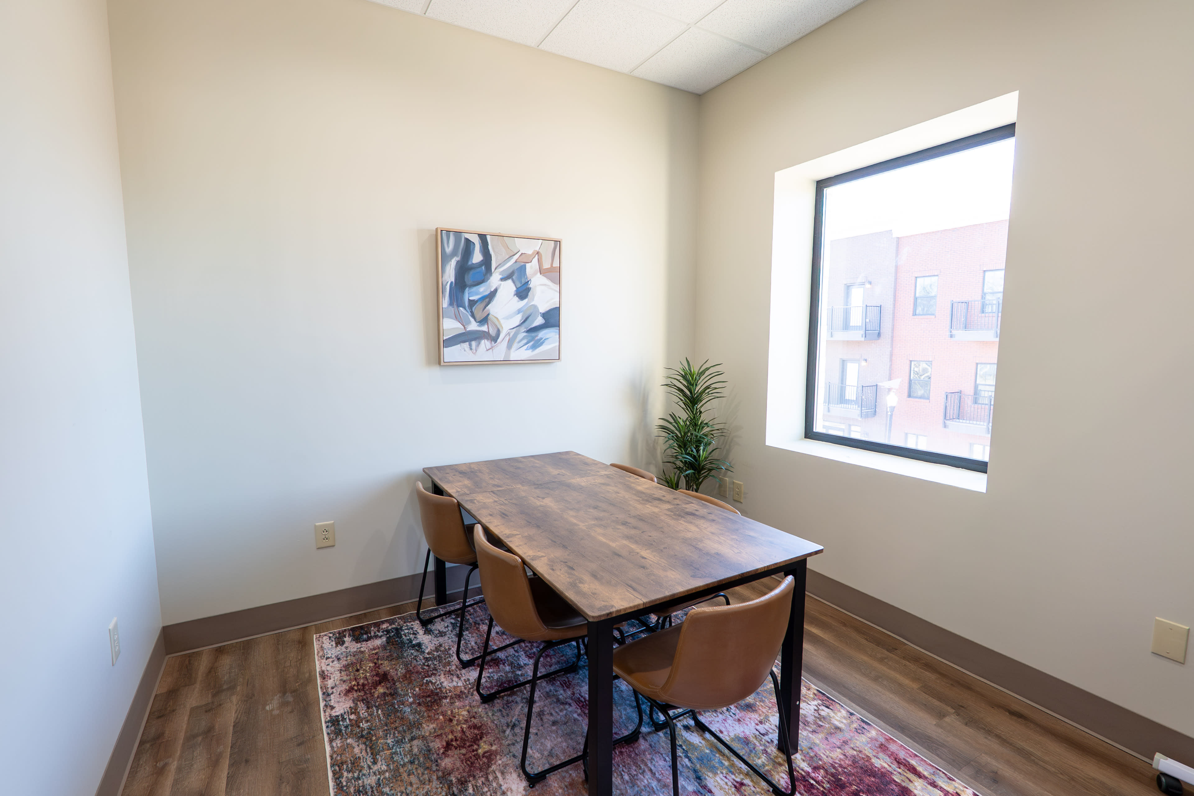 The image shows a small meeting room with a wooden table and four chairs, a decorative plant, and a window.