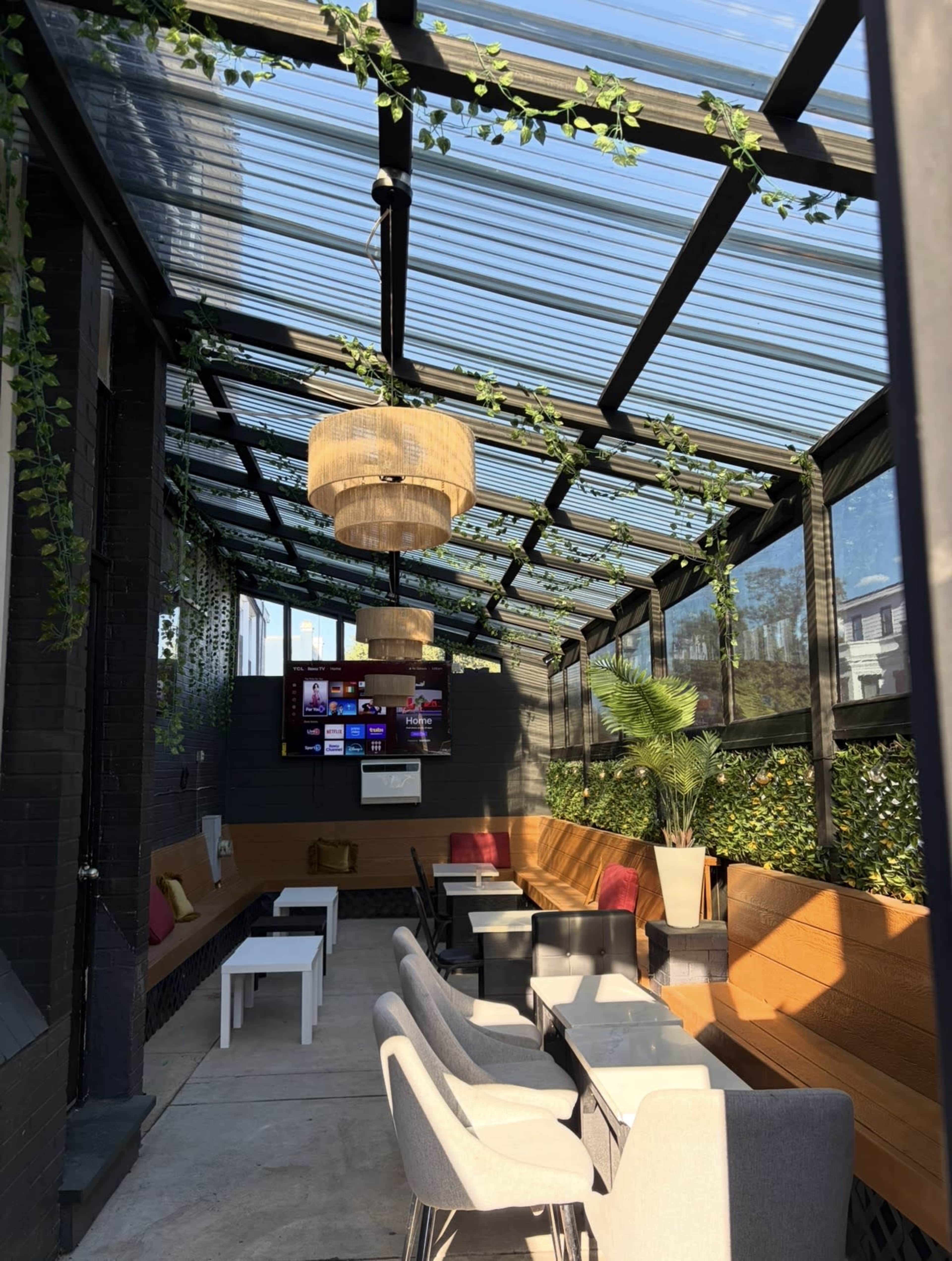 The image shows a modern outdoor terrace area with glass roofing, wooden seating, and decorative plants.
