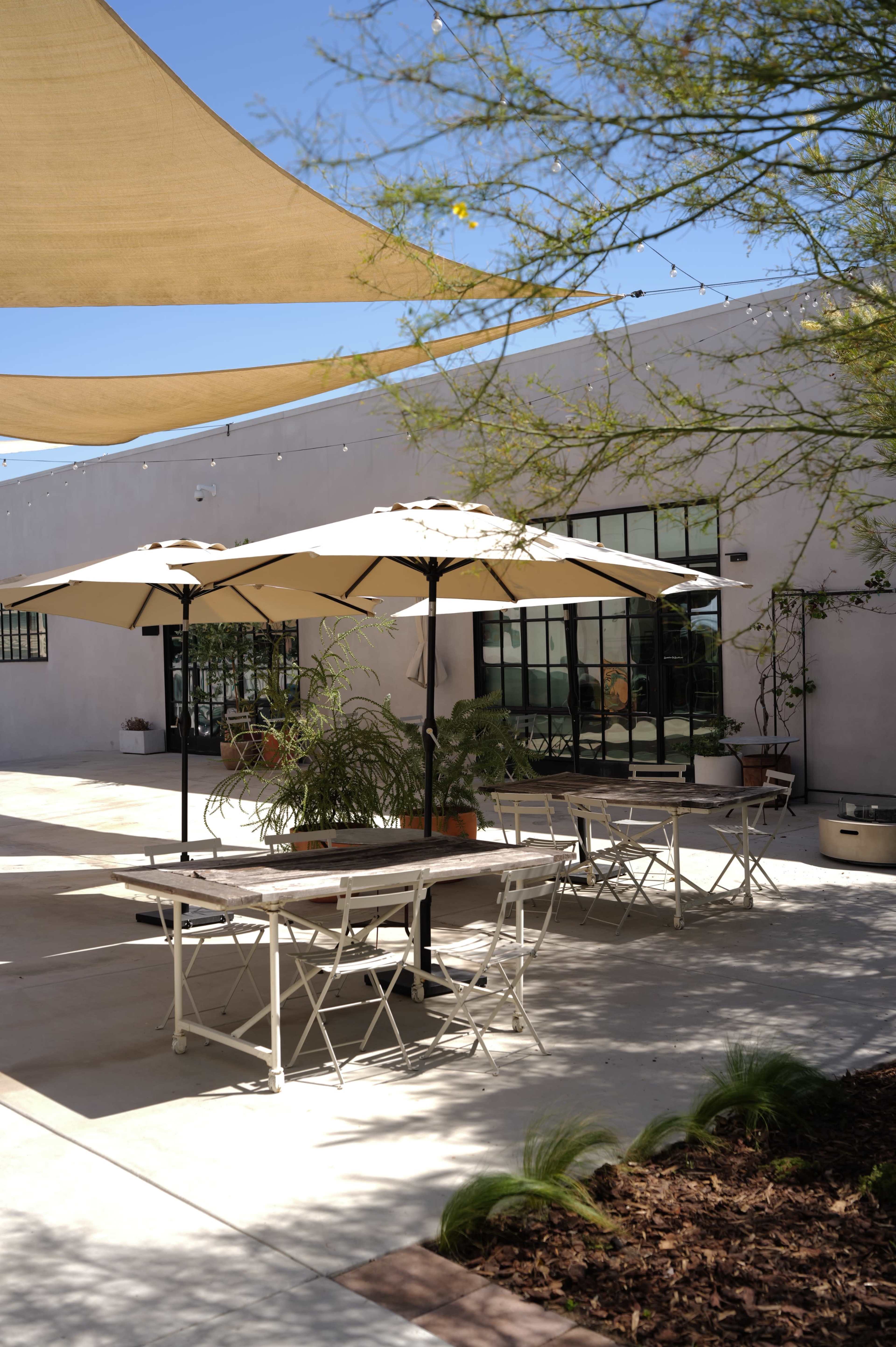 A patio area features multiple tables with umbrellas, surrounded by plants and modern architectural elements under a blue sky.