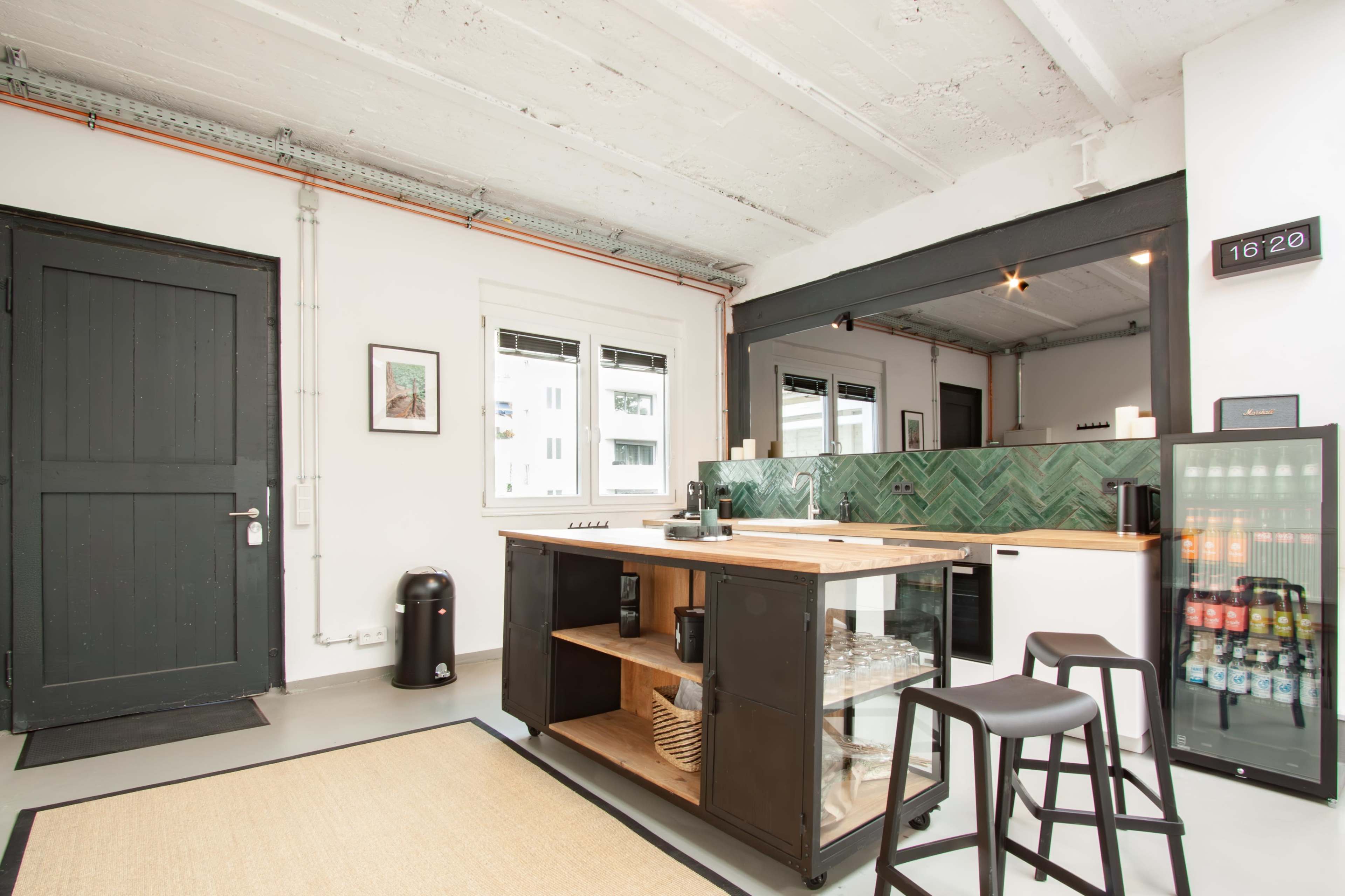 A modern kitchen features a central island with a wooden countertop, black shelves, and a green tile backsplash, alongside a black trash can and a small fridge.