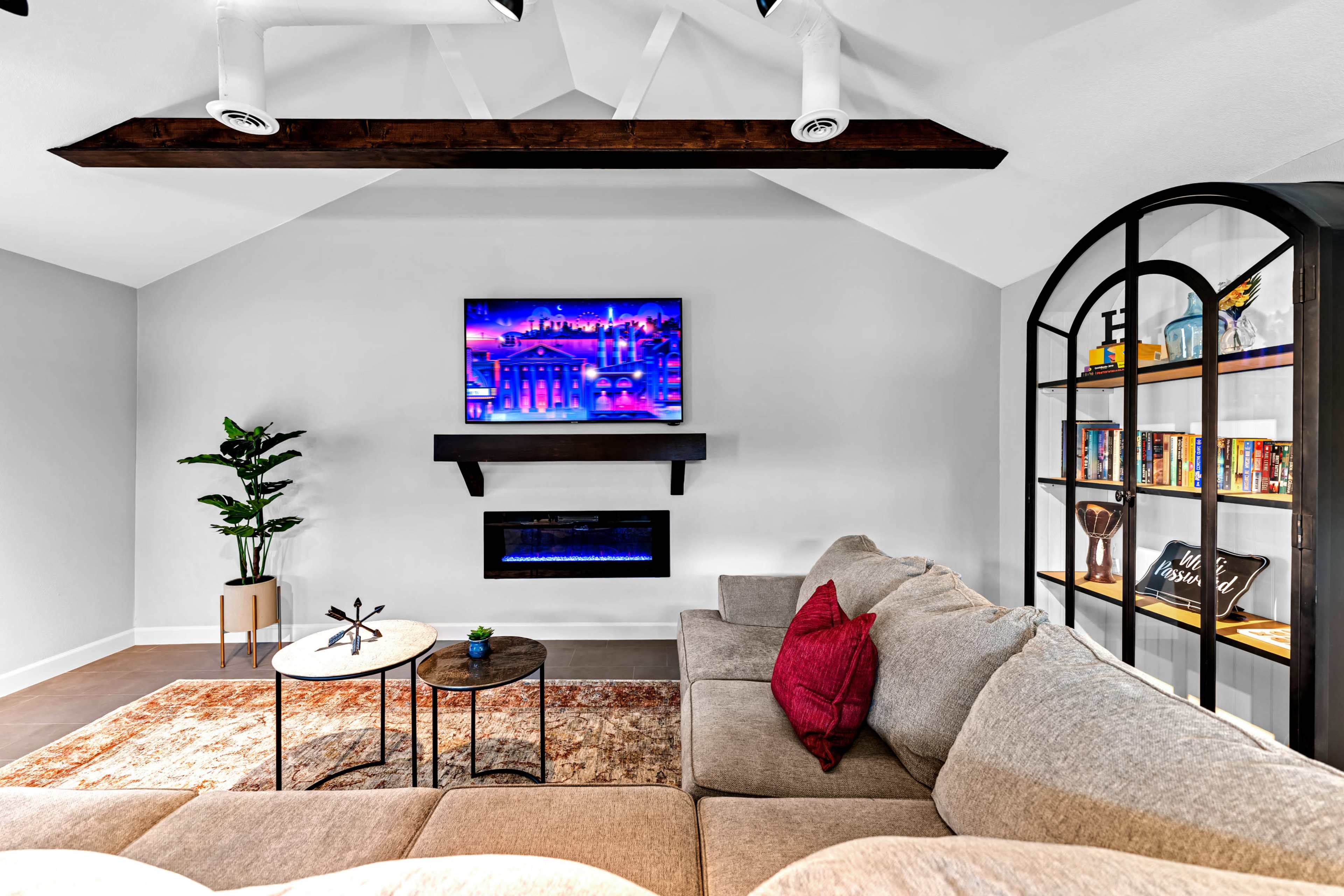 A modern living room features a sectional sofa, a wall-mounted television, an electric fireplace, and a bookshelf filled with books, all set against a vaulted ceiling with exposed beams.