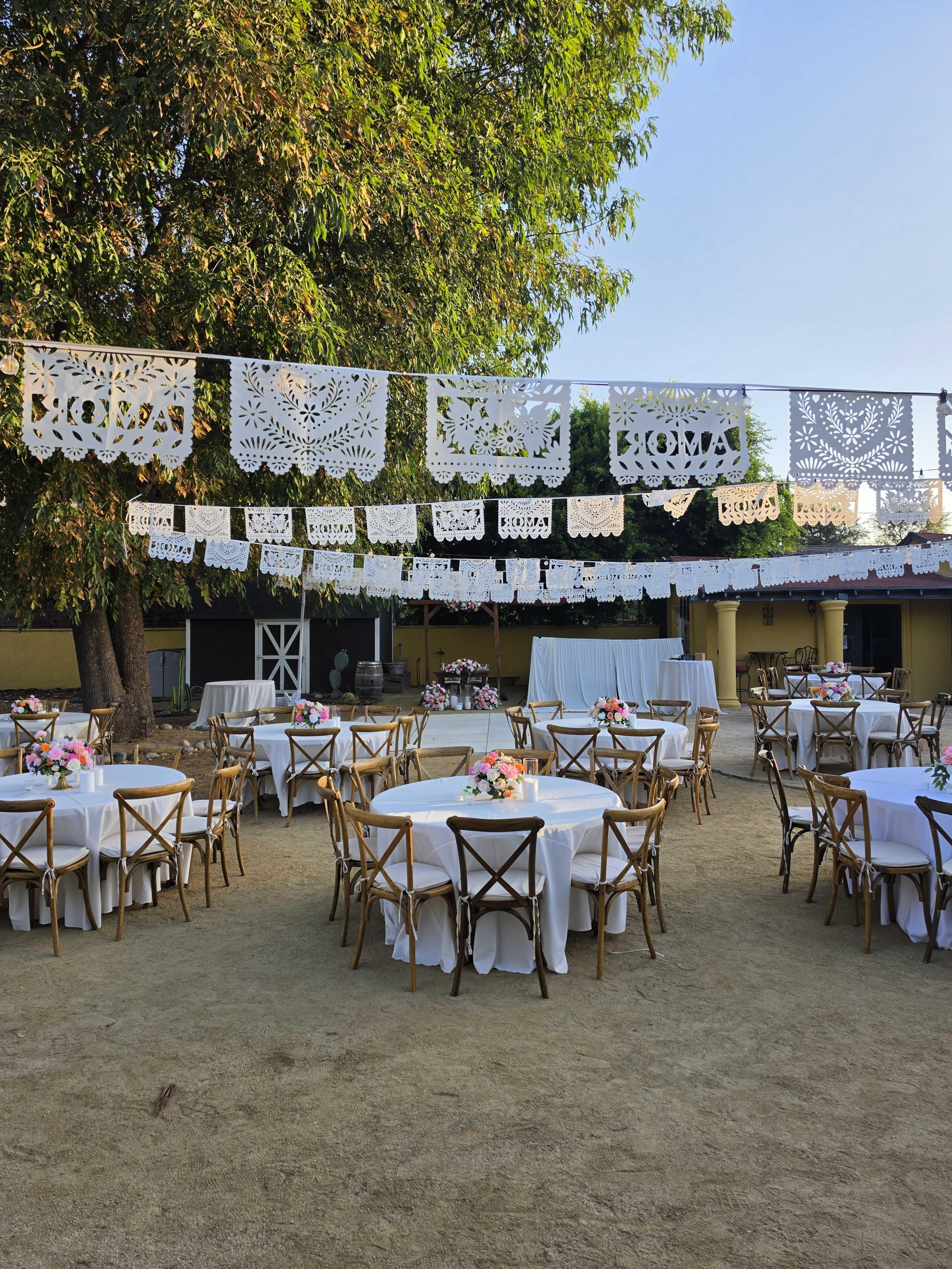 The image shows a decorated outdoor event space with round tables covered in white linens, surrounded by rustic wooden chairs, and adorned with floral centerpieces under colorful papel picado banners.