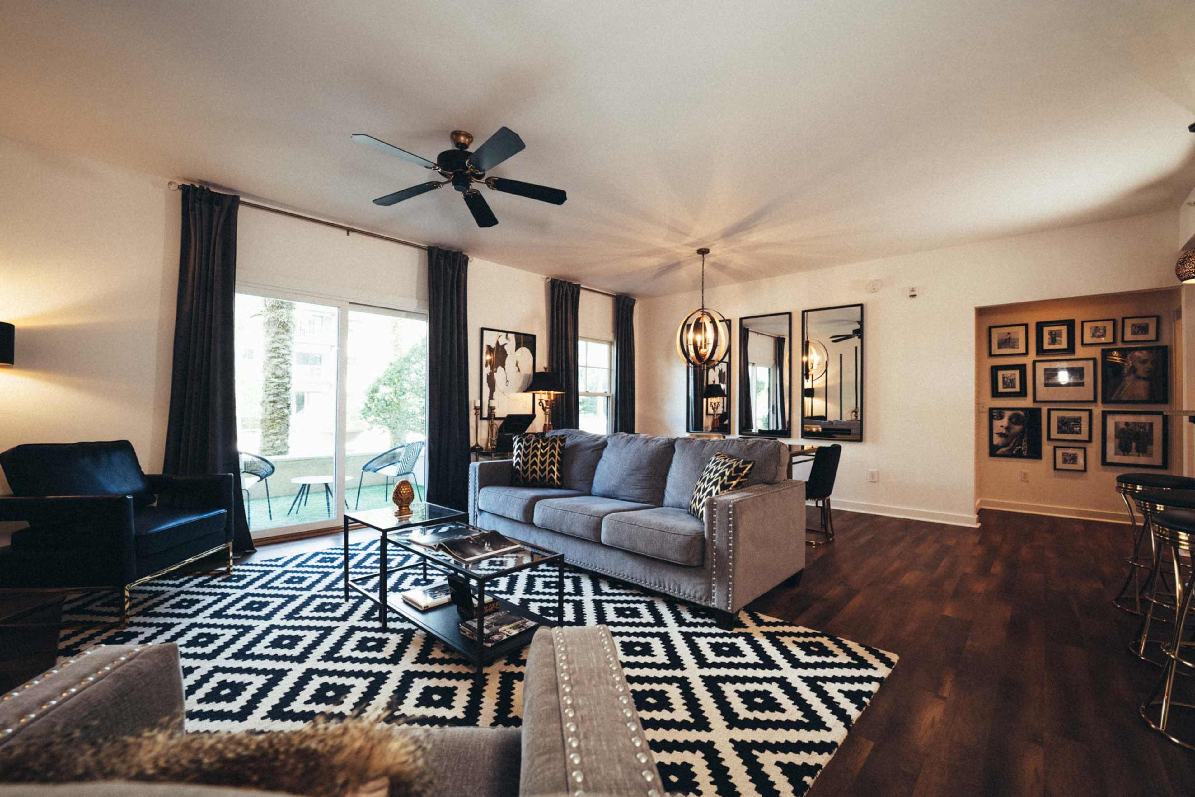 The living room features a gray sofa, a patterned rug, and large windows with dark curtains, complemented by a dining area with framed photographs on the wall.