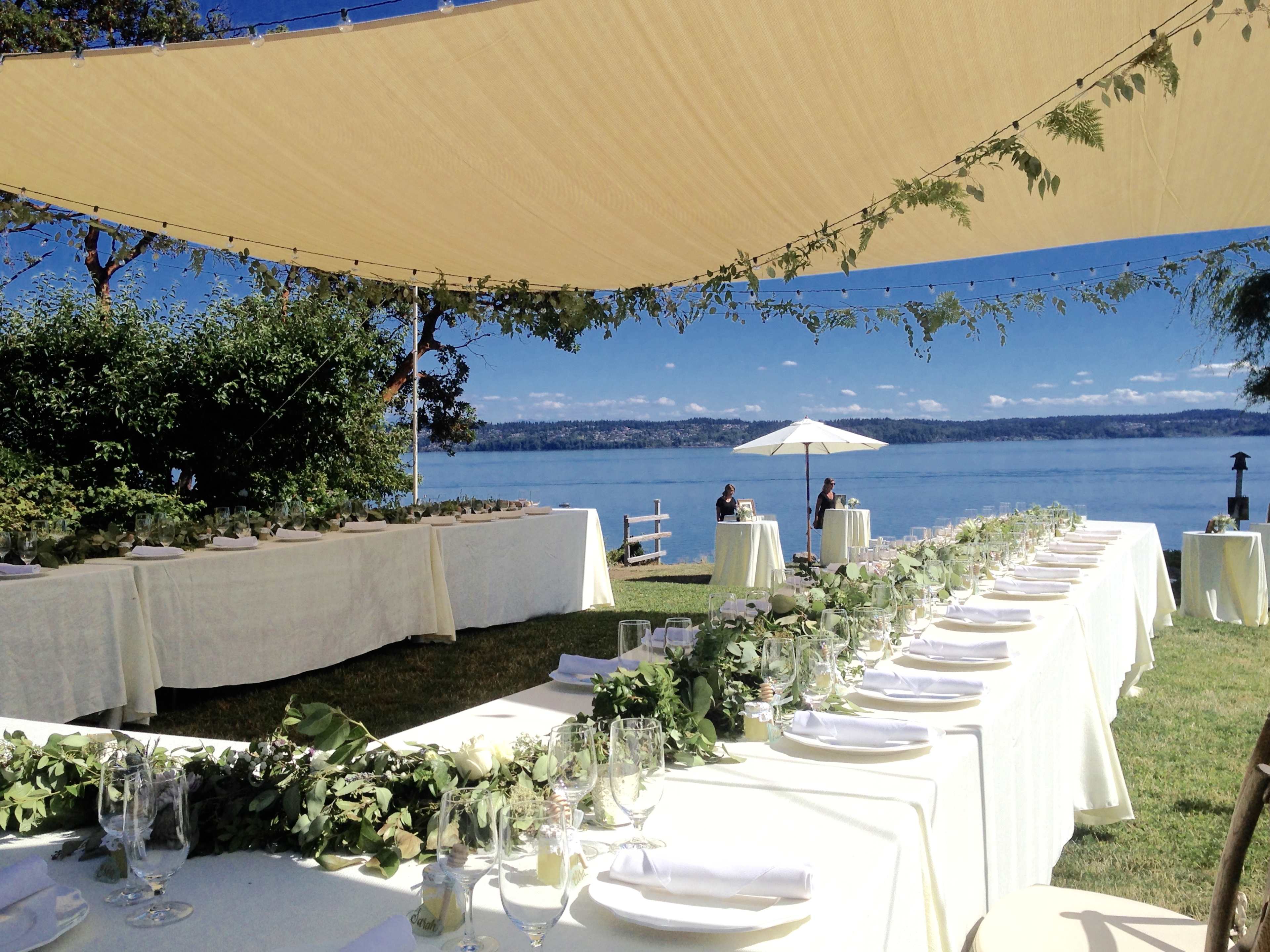 A long, elegantly set dining table is arranged under a shaded canopy beside a calm waterfront, with greenery and white tableware.