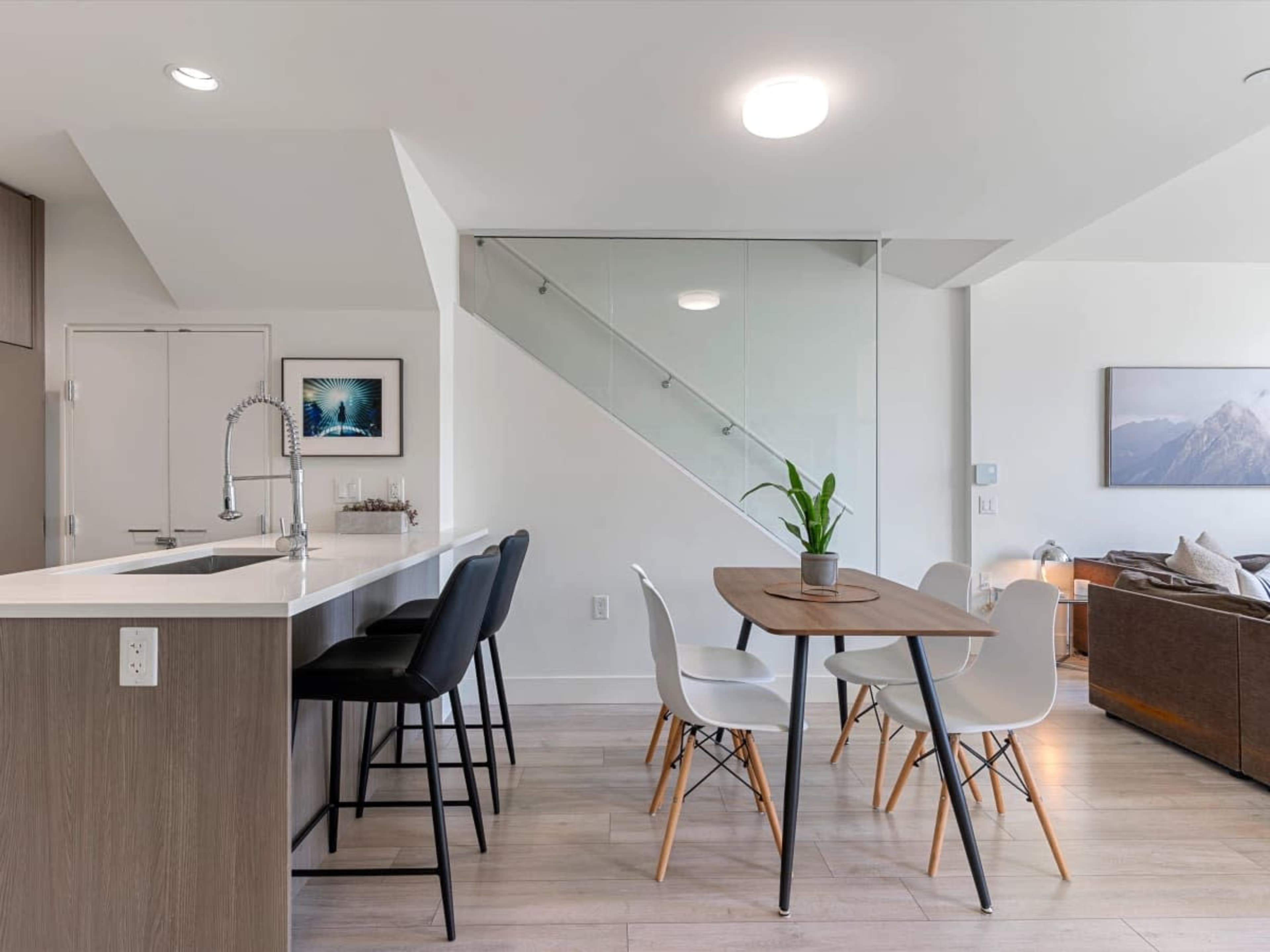 A modern open-concept living space features a kitchen bar, a dining table with four chairs, and a staircase with glass railings.