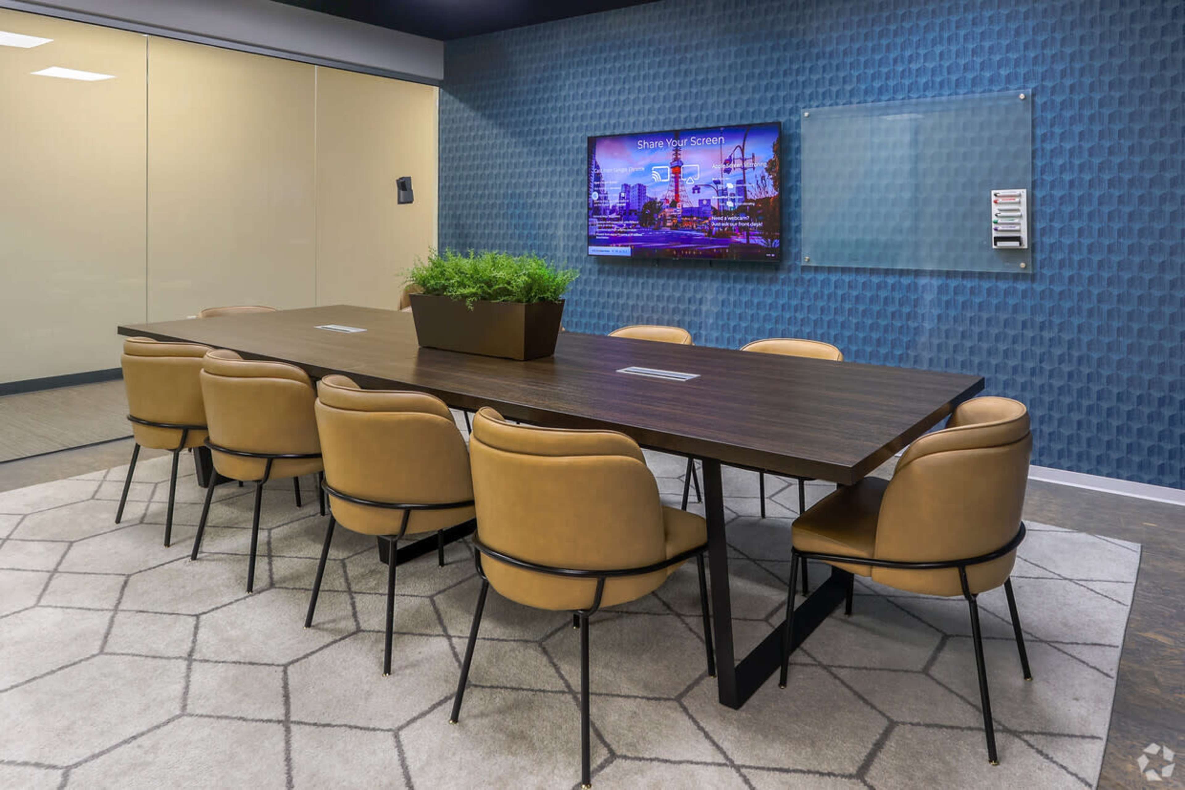 The image shows a modern conference room with a long wooden table surrounded by eight brown chairs, a television mounted on the wall, and a potted plant as a centerpiece.