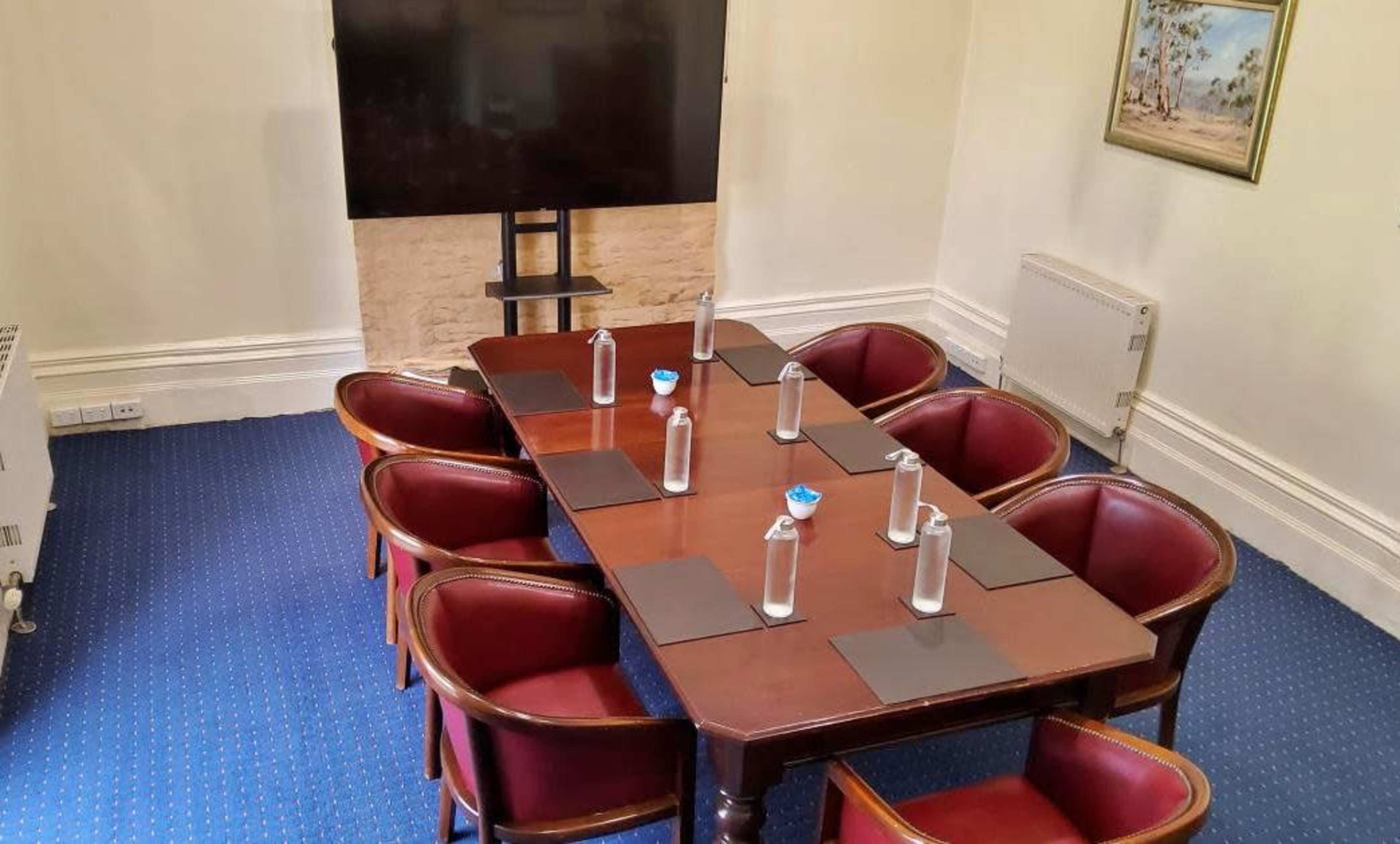 A conference room features a rectangular wooden table surrounded by eight chairs, with a large screen mounted on the wall and water bottles on the table.