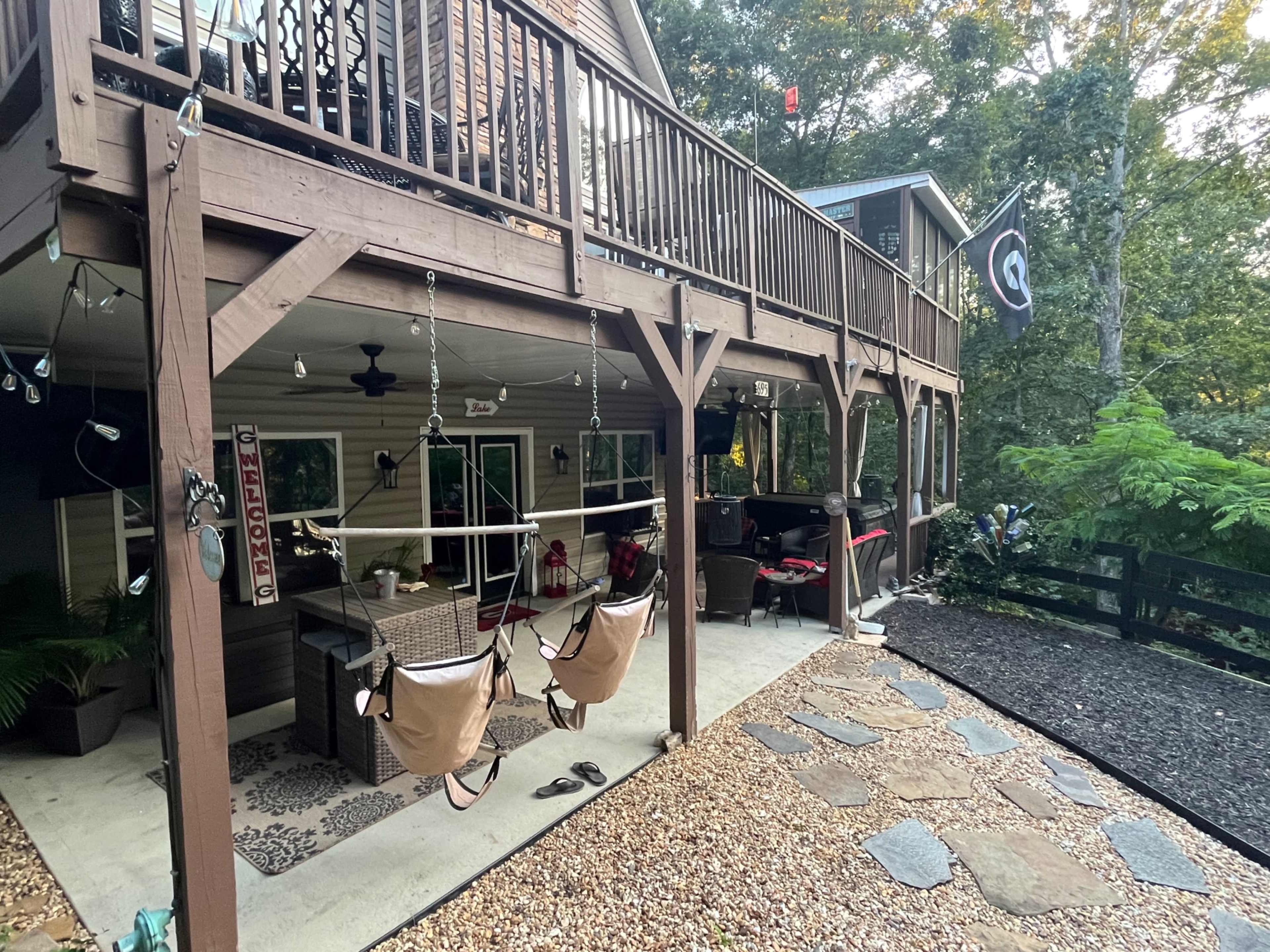 The image shows a two-story wooden deck with a seating area and hanging chairs, surrounded by trees and a gravel pathway.
