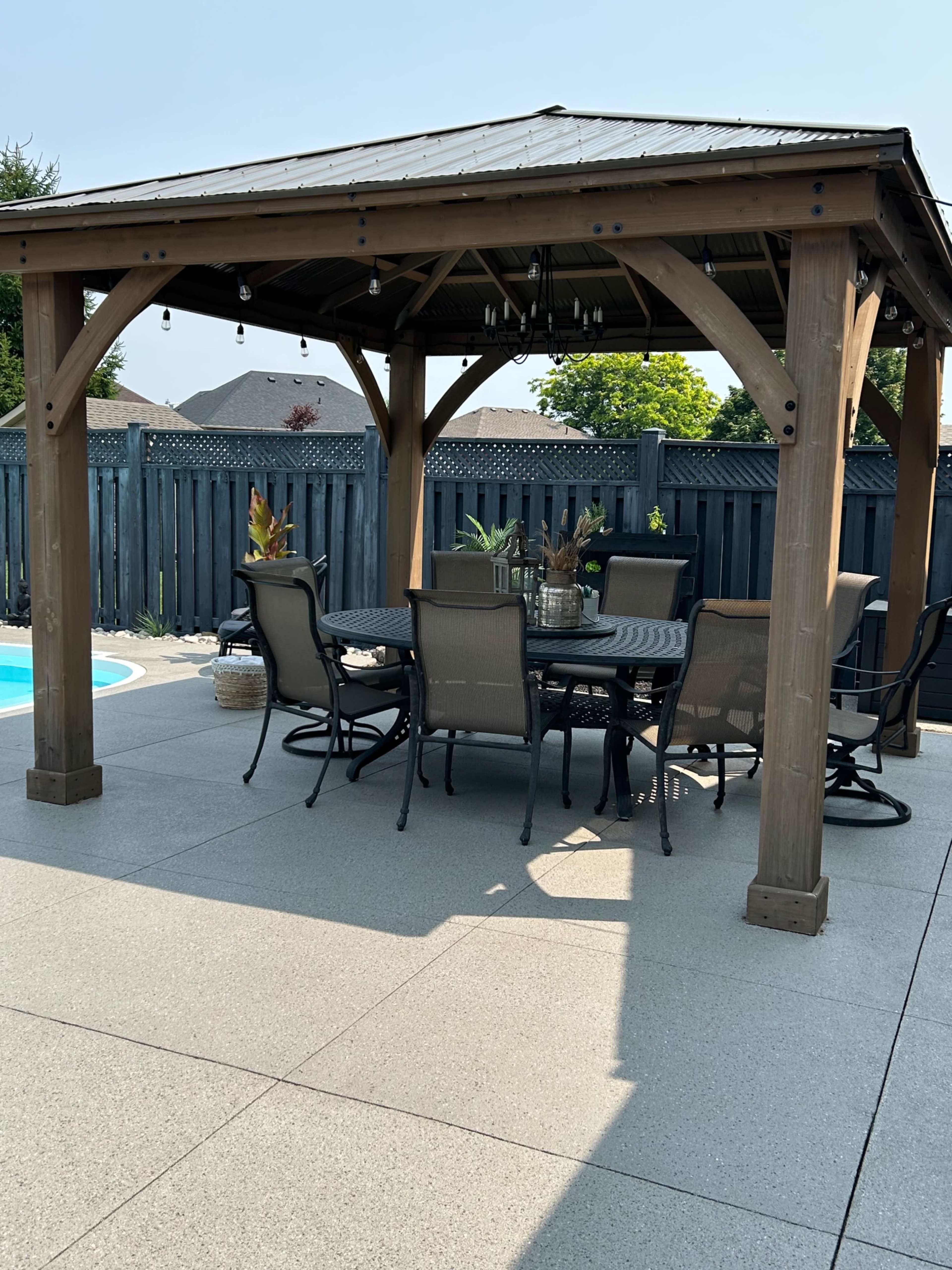 A gazebo with a round table and chairs is set on a concrete patio beside a swimming pool, surrounded by a wooden fence and greenery.