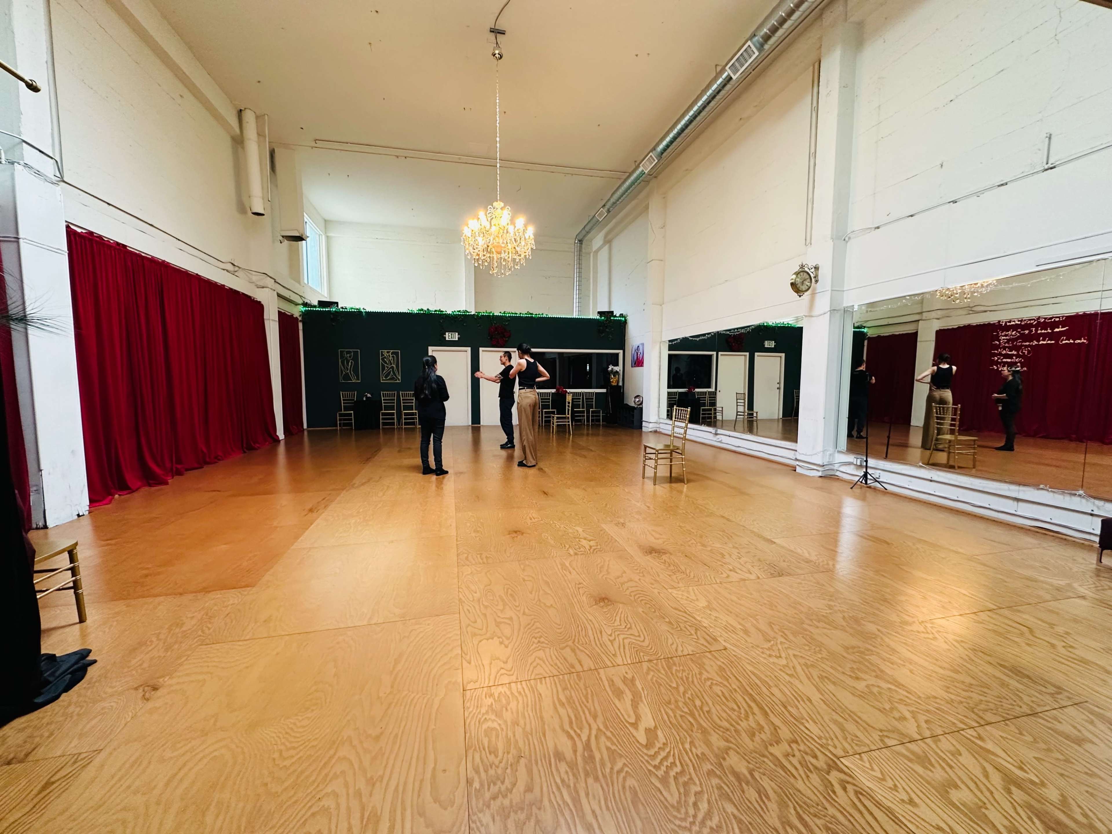 A spacious room with wooden flooring, red curtains, mirrors on one wall, and two people engaged in conversation.