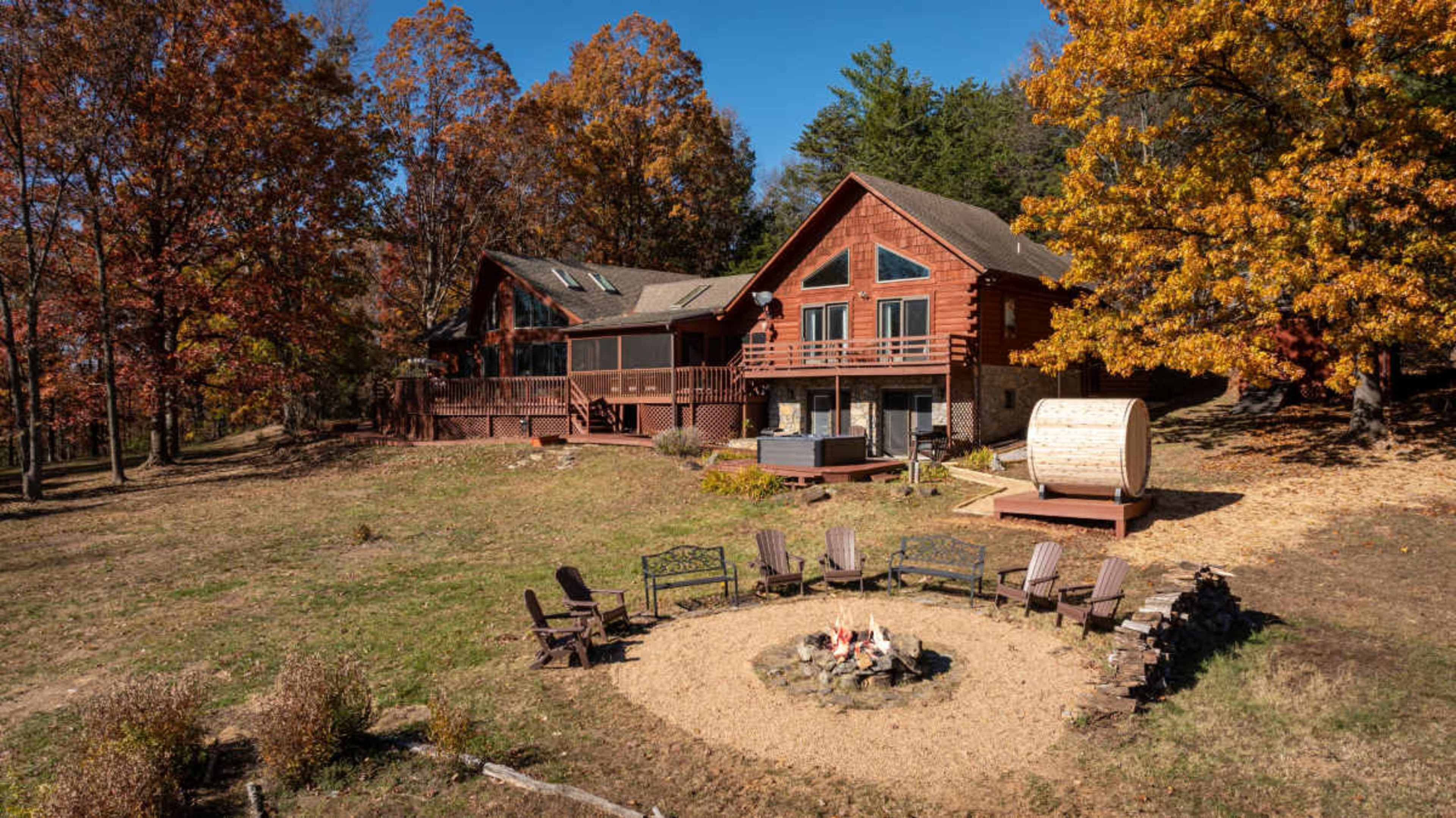 A large wooden house with multiple levels and a wraparound deck is surrounded by colorful autumn trees and a circular fire pit with seating.