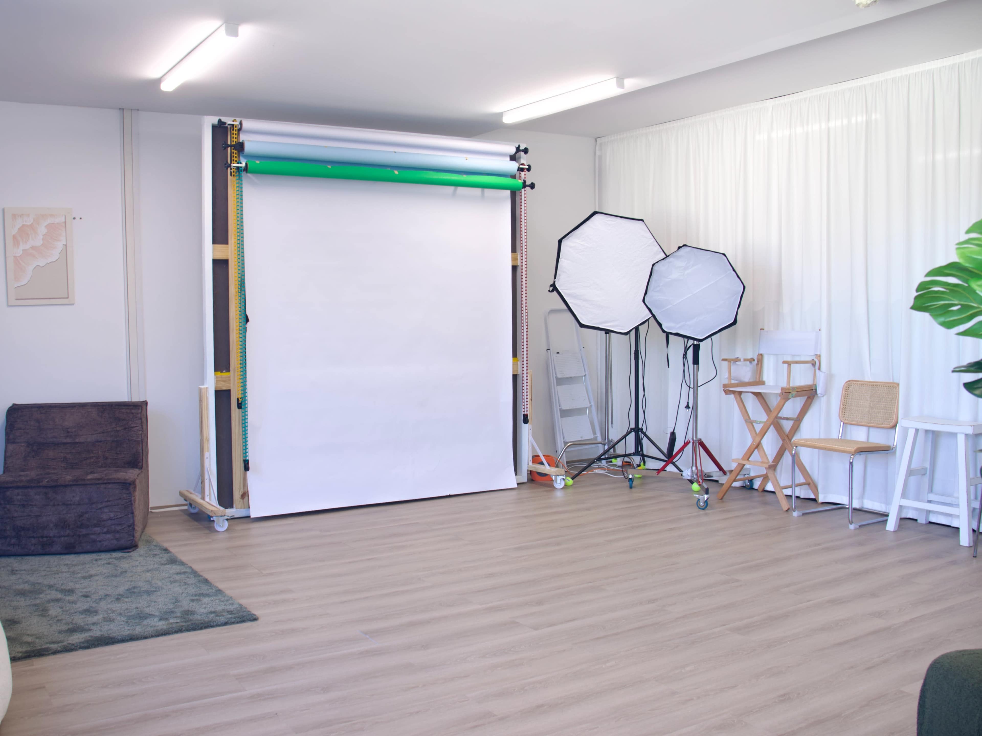 The image shows a photography studio setup with a backdrop, lighting equipment, and various seating arrangements against a light-colored wall.
