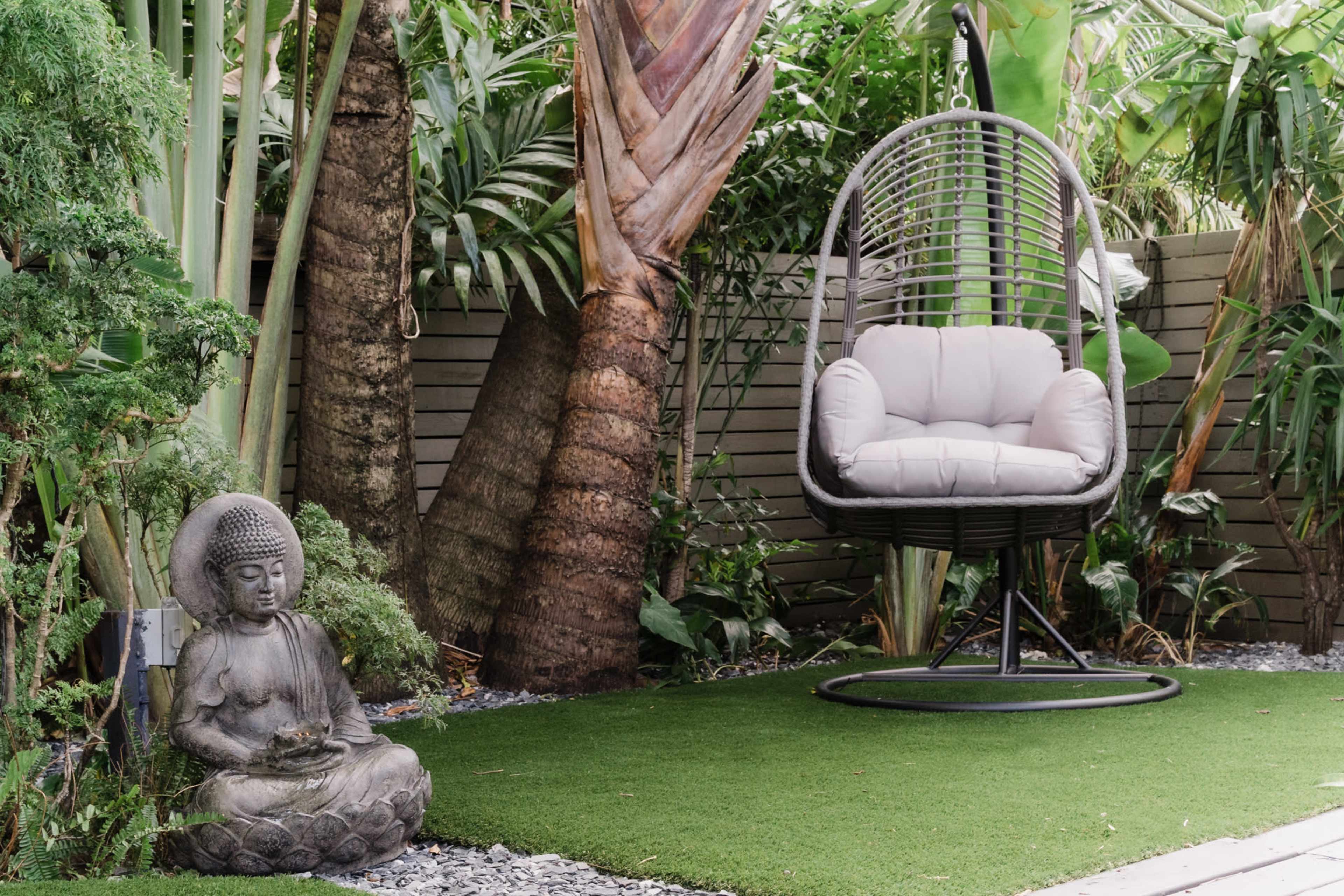 A serene garden scene with a hanging chair and a stone Buddha statue surrounded by lush greenery and artificial grass.