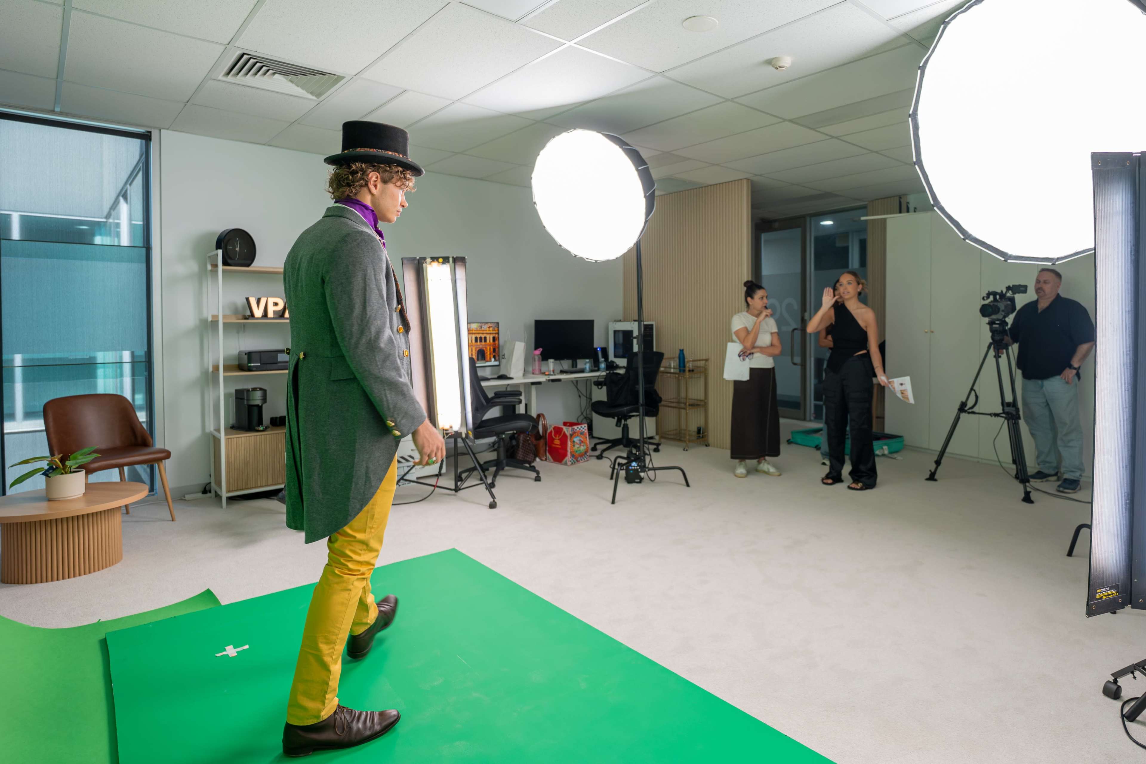 A man wearing a top hat and colorful outfit walks on a green screen in a well-lit office space while a small group observes and records.